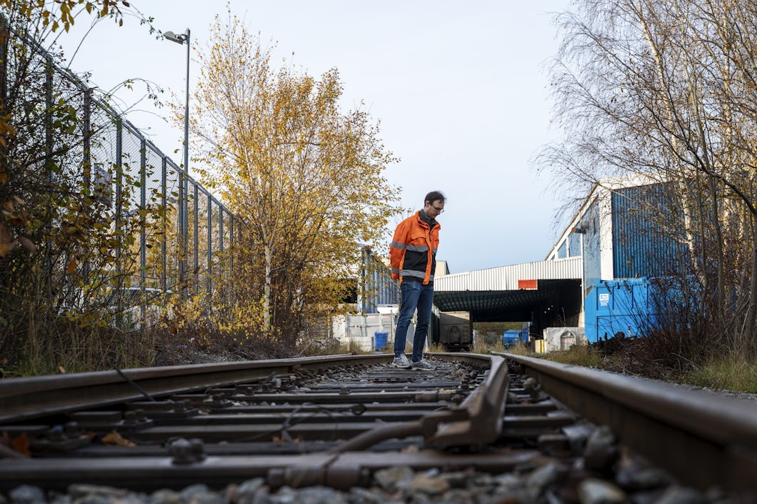 Fredrik Stangel med orange jacka undersöker ett böjt järnvägsspår nära industribyggnader och träd med höstlöv.