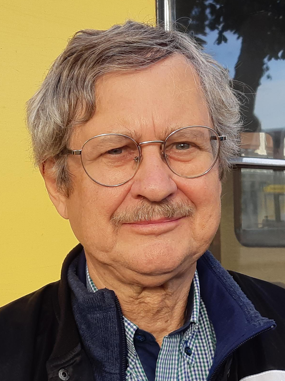 Bo-Lennart Nelldal med grått hår, glasögon och mustasch, har skjorta med krage och jacka.