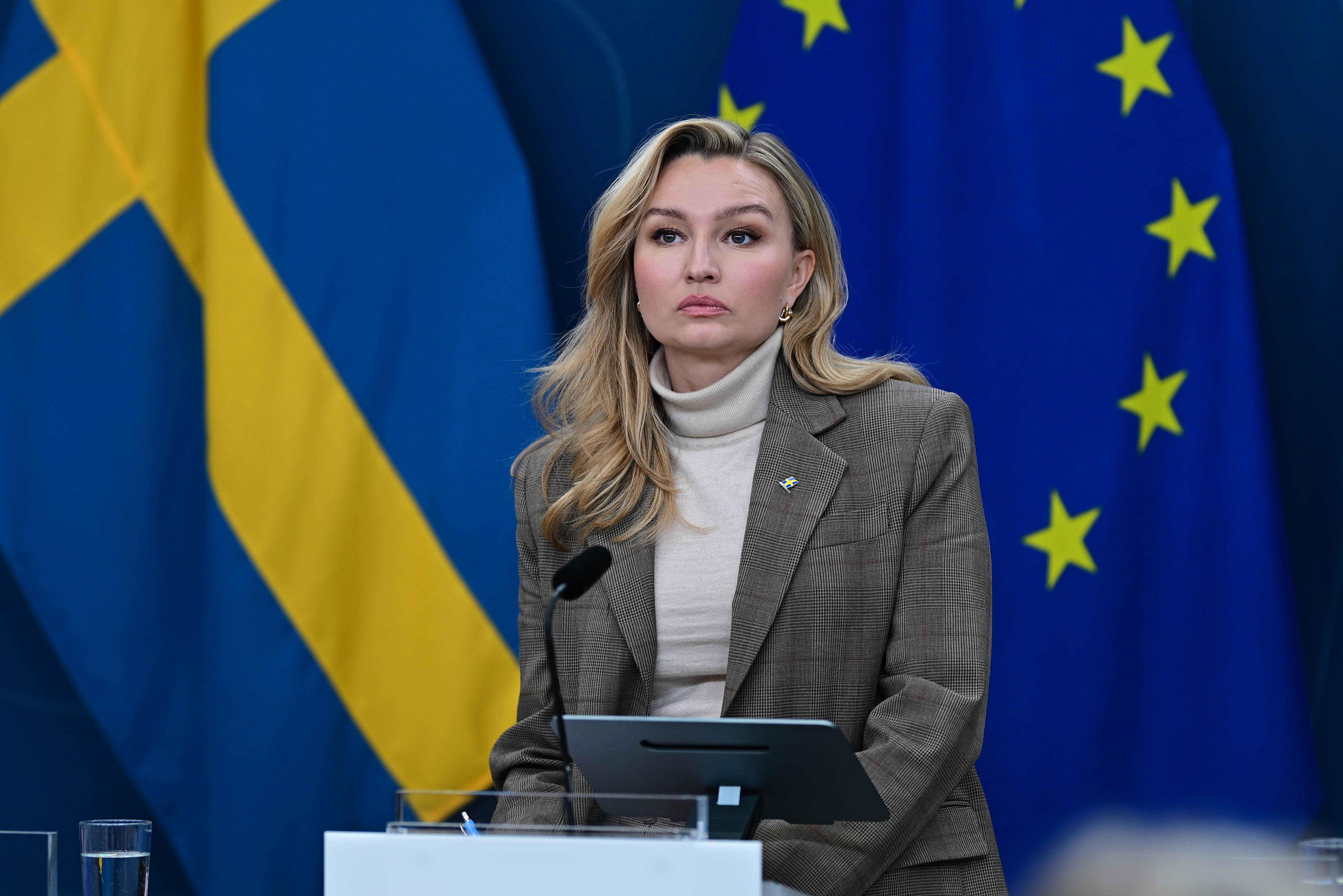 En kvinna i beige kostym står vid ett podium med mikrofoner framför svenska och europeiska unionens flaggor.