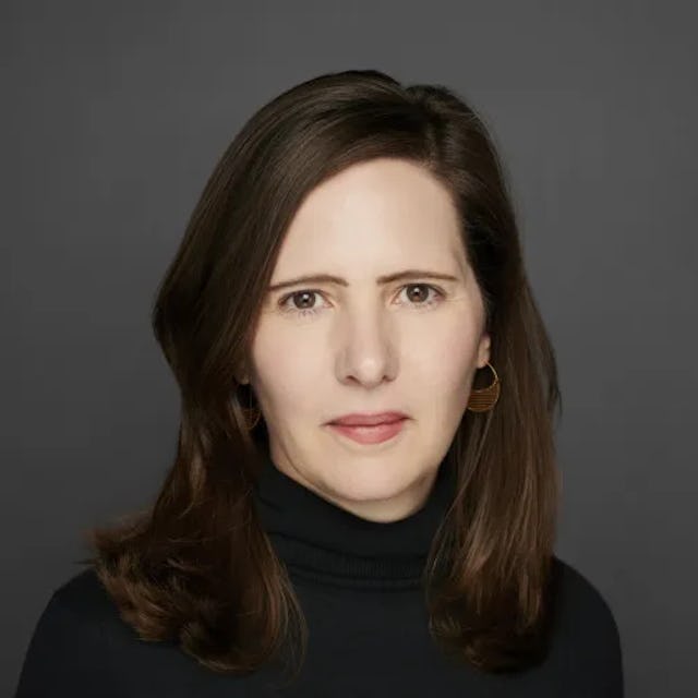 En kvinna med rakt brunt hår, en svart pikétröja och guldörhängen står på en mörkgrå bakgrund.