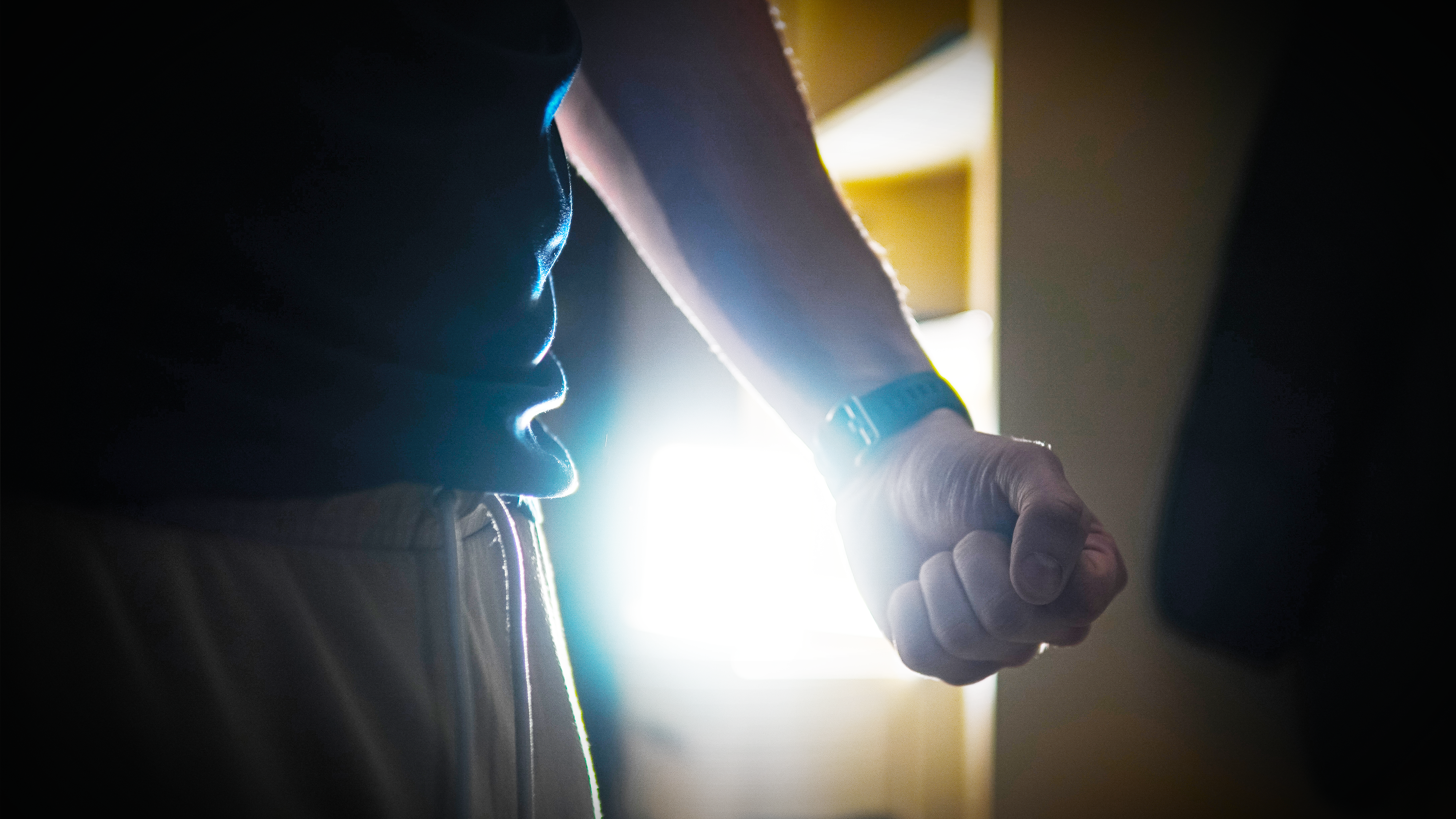 En person står inomhus med knuten hand, med starkt ljus bakom sig.