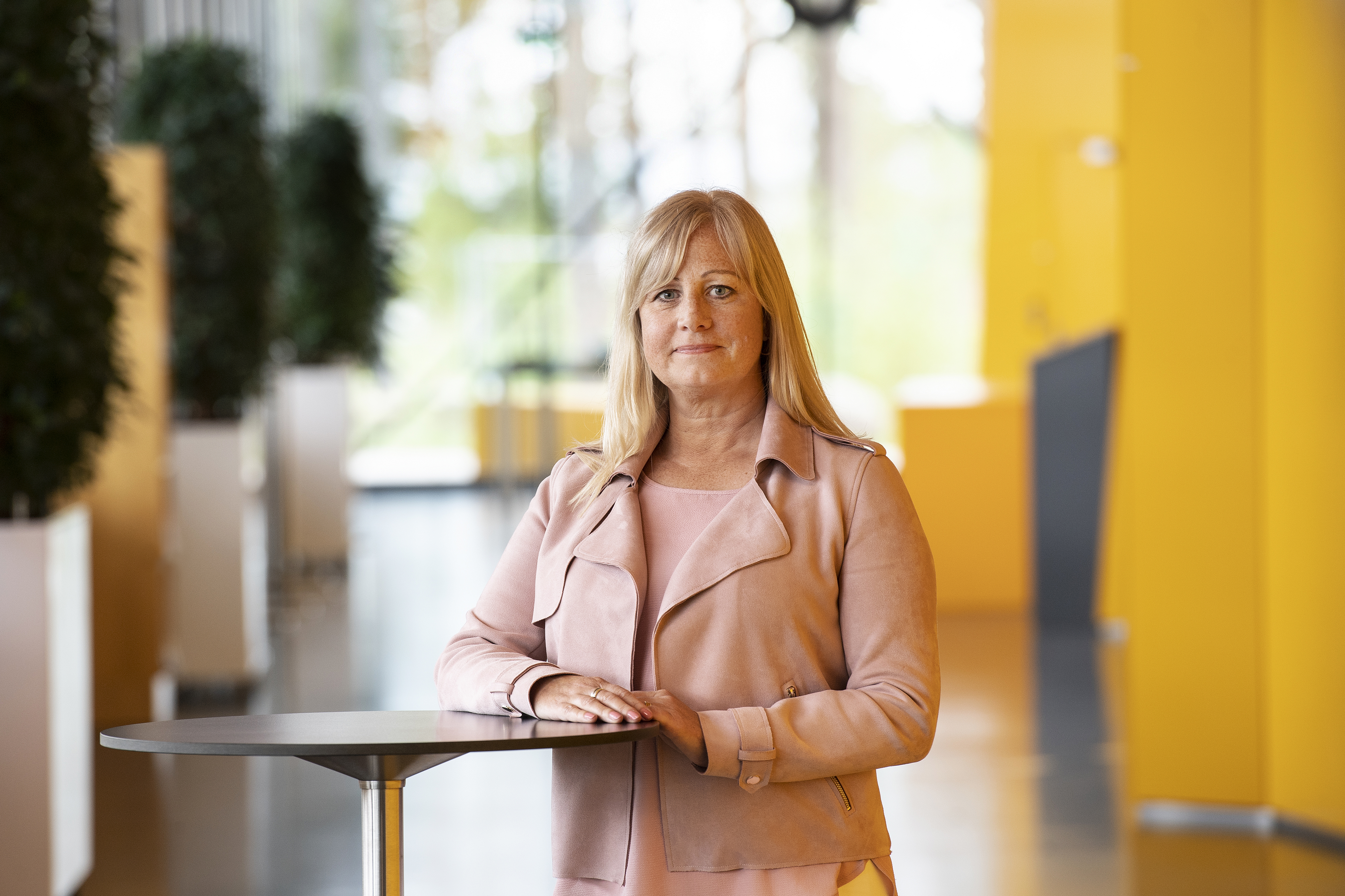 En kvinna med blont hår och en ljusrosa jacka står vid ett runt bord i ett inomhusutrymme med gula väggar och gröna växter.