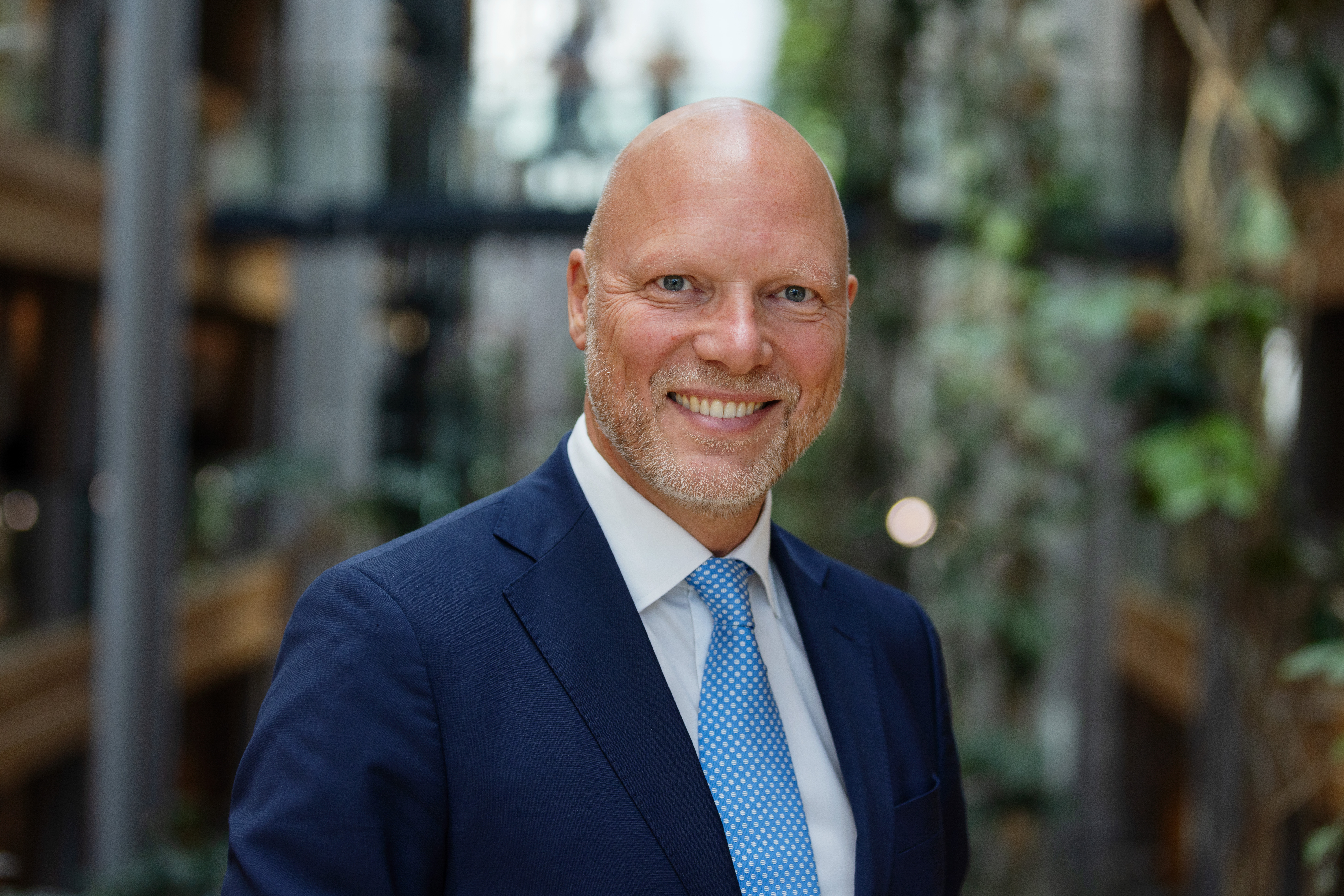 En man i blå kostym och ljusblå slipsar står inomhus med suddiga växter och byggnadsdetaljer i bakgrunden.