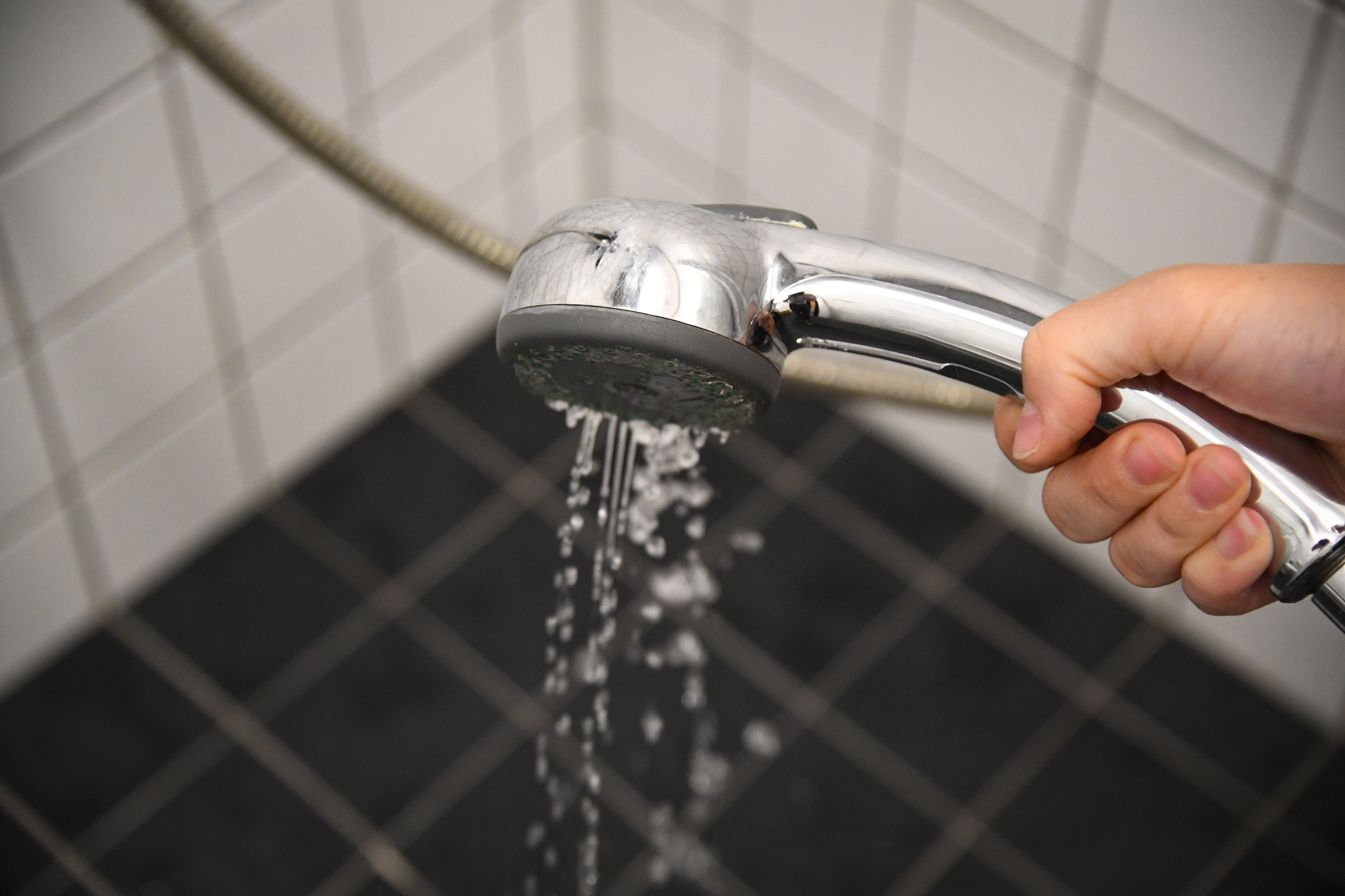 En hand håller en duschslang med vatten som rinner, i ett hörnduschutrymme med vita väggar och mörka golvplattor.