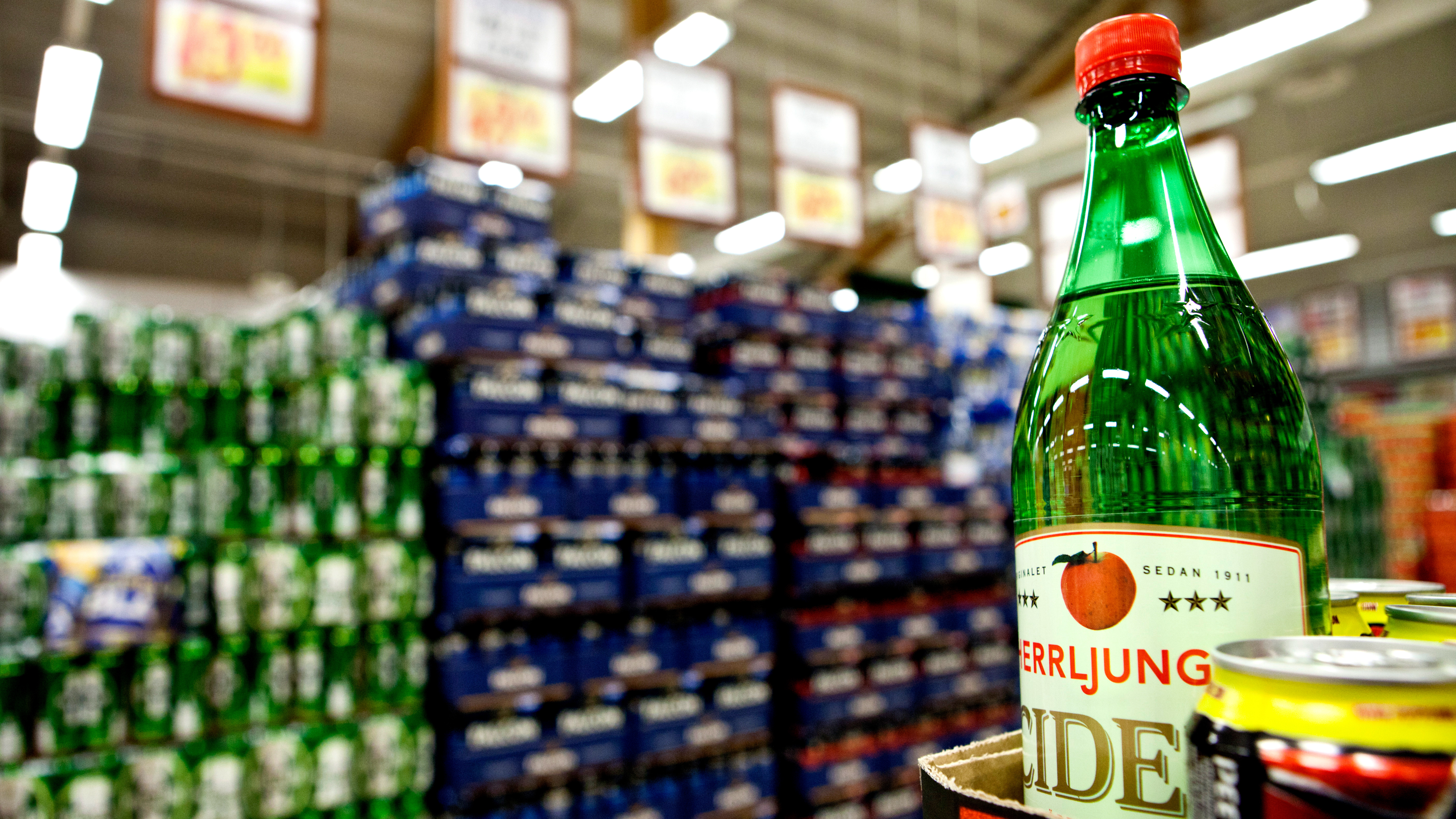 En närbild av en grön glasflaska med cider på en butikshylla, med staplade dryckeslådor i bakgrunden.
