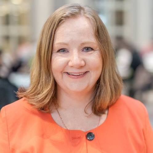 En kvinna med axellångt ljusbrunt hår, iförd orange kavaj, ler mot kameran i en inomhusmiljö med suddig bakgrund.