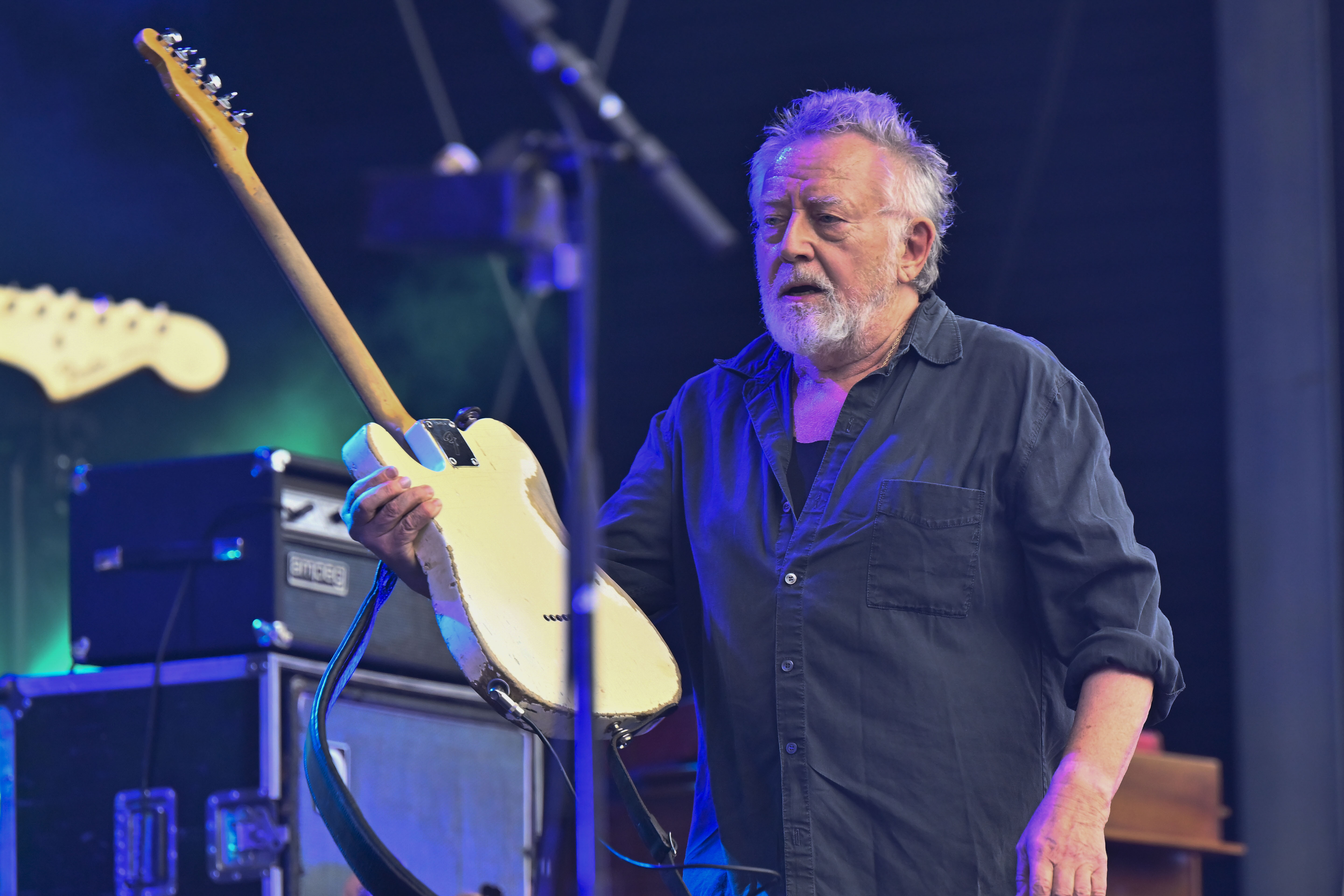 En äldre man med grått hår och skägg står på en scen och håller i en elgitarr, med ljudutrustning i bakgrunden.