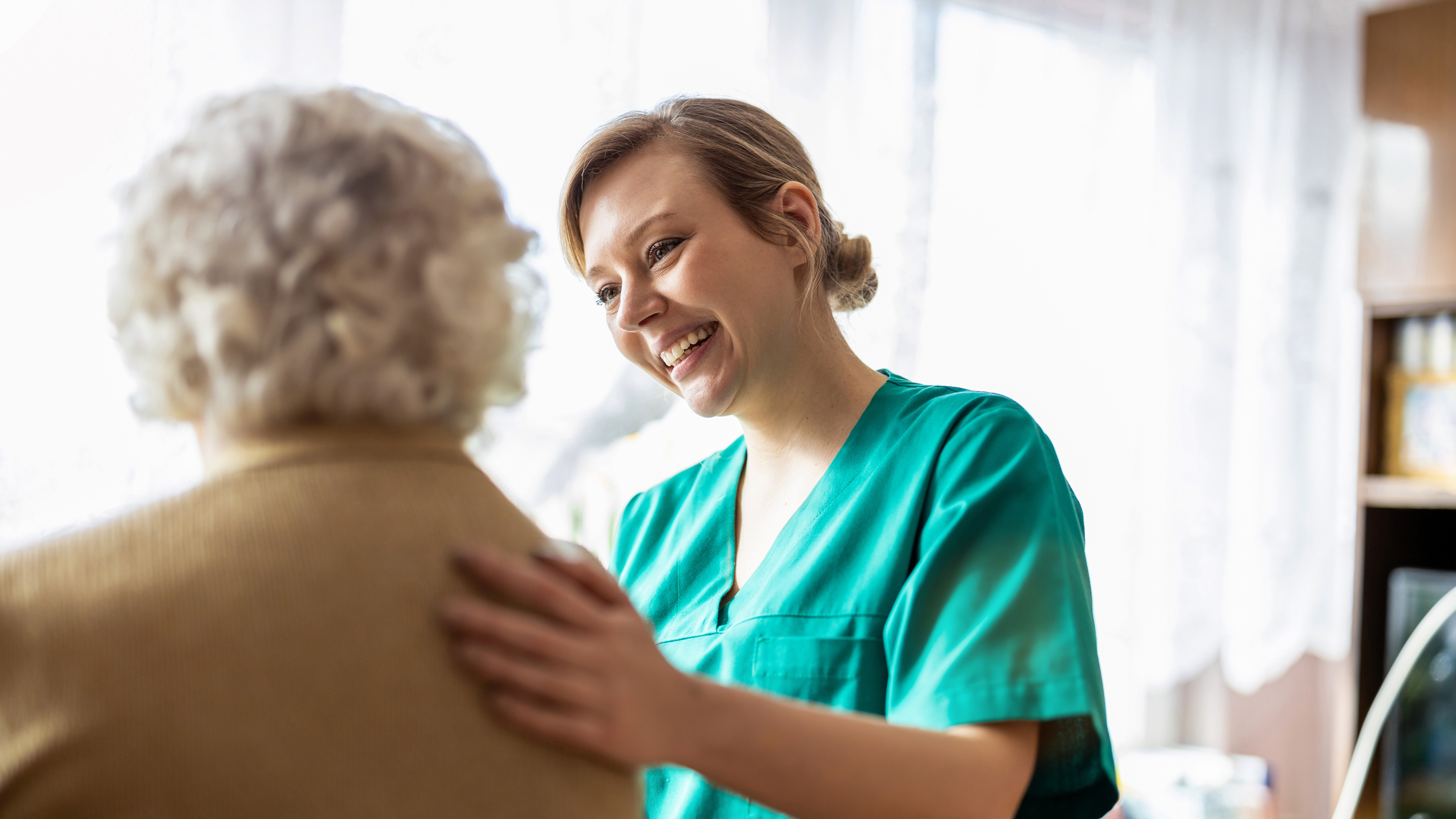 En vårdare i gröna arbetskläder ler och lägger sin hand på axeln av en äldre man med grått hår.