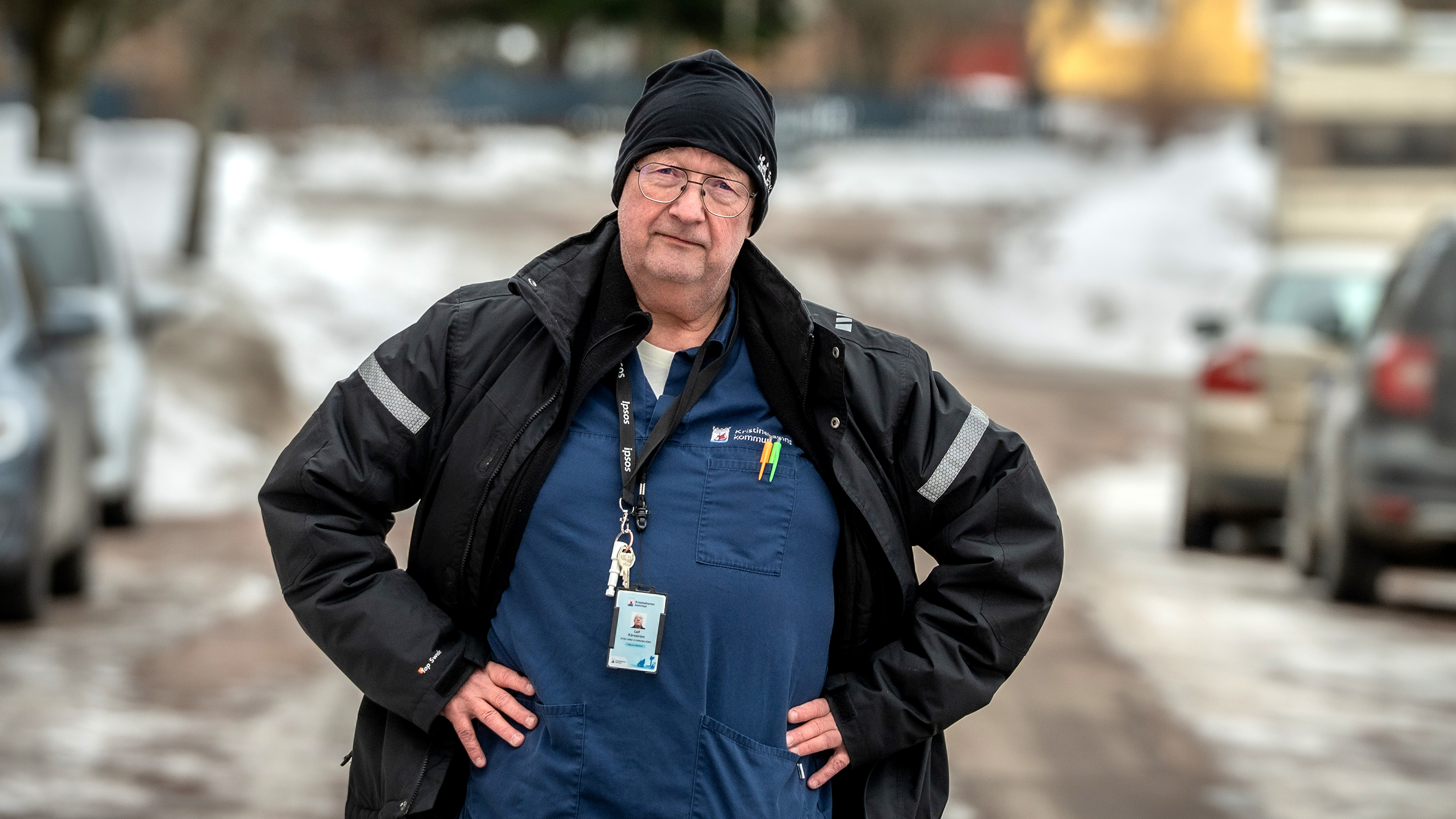 En äldre person med glasögon, vintermössa och jacka står med händerna på höfterna på en snötäckt gata, har namnbricka och flera pennor i skjortfickan.