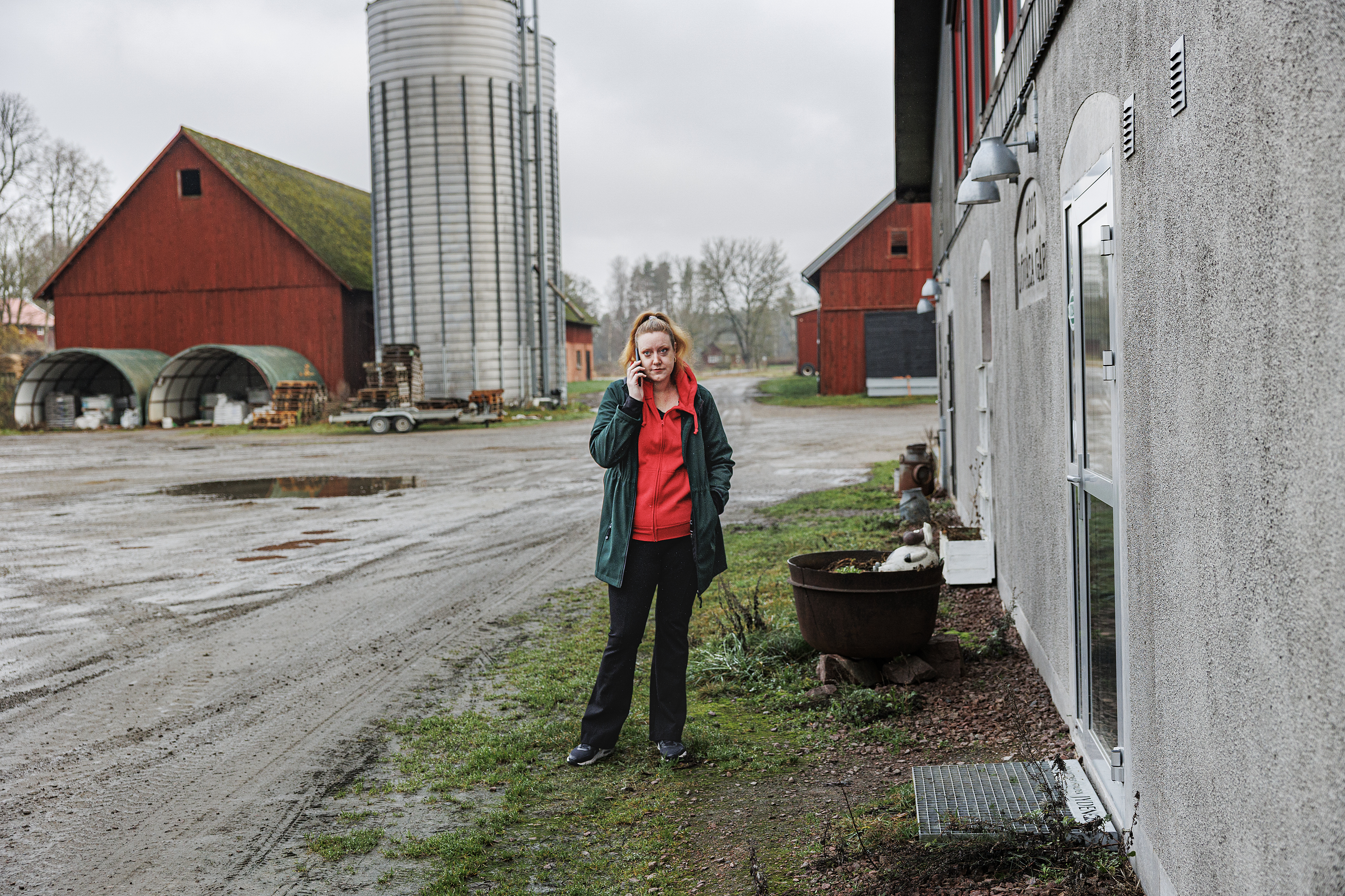 En person står utanför en ladugård och pratar i mobil. I bakgrunden syns lador, en silo och utrustning.