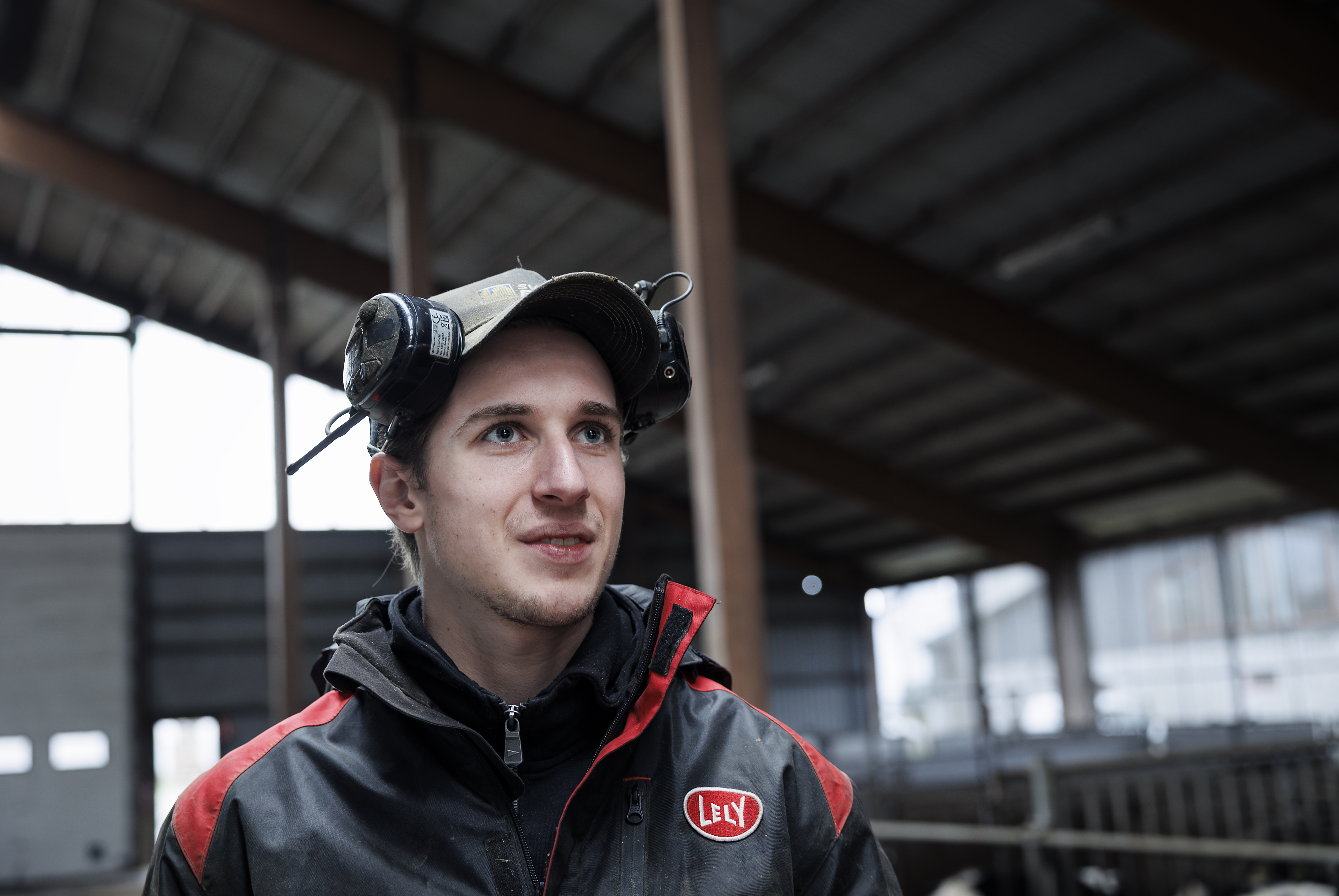 En man iklädd jacka och hatt med hörselskydd står i en lada med metallbalkar och djurlådor i bakgrunden.