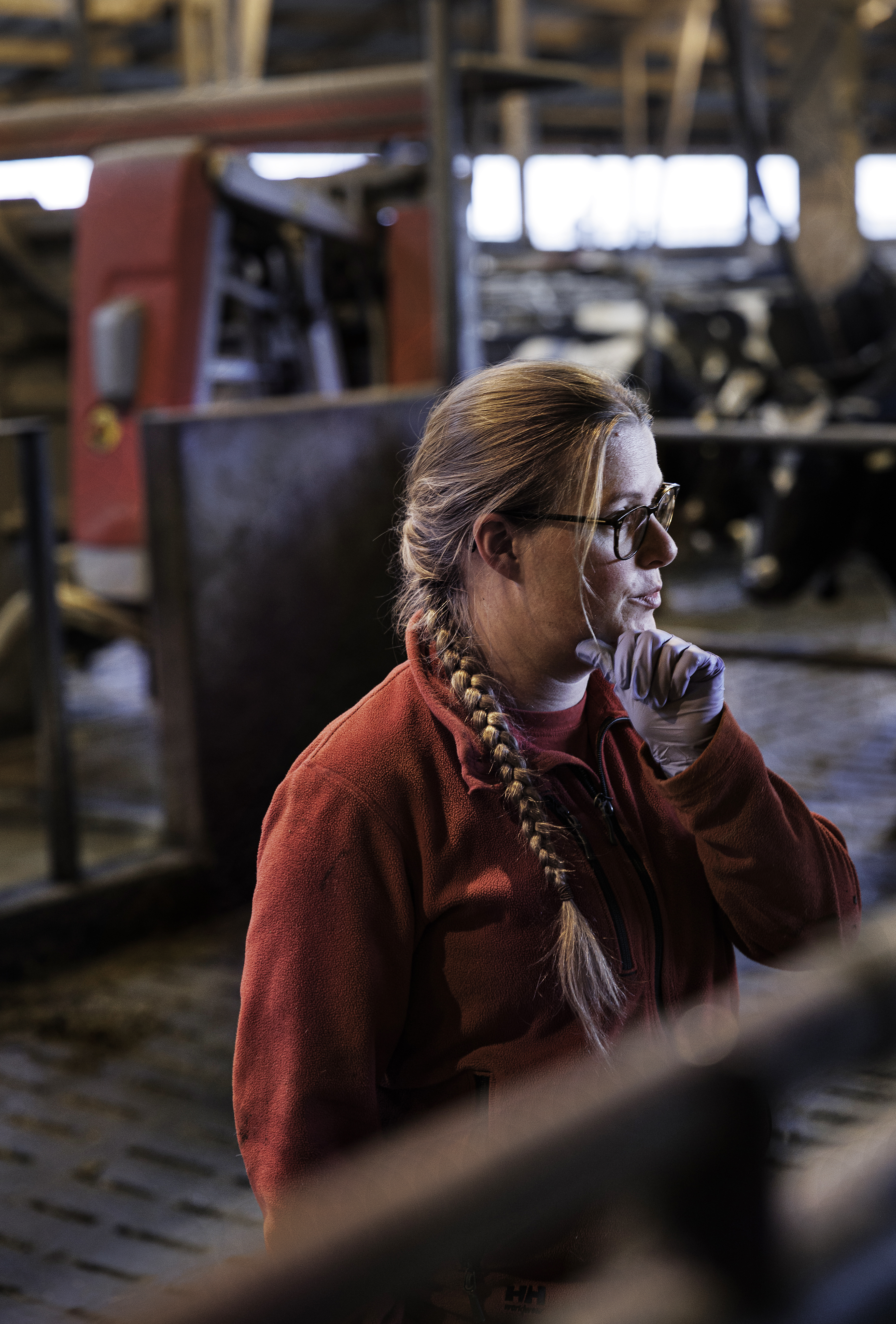 En kvinna med långa flätor, glasögon och en röd jacka står vid en boskapsbod inomhus. Kor och jordbruksutrustning kan ses i bakgrunden.