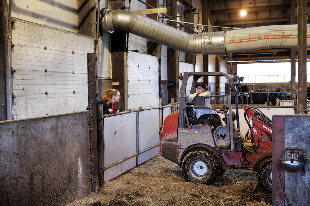 En person kör en liten lastare i ladugården och den andra står vid grinden, omgiven av jordbruksutrustning och hö.