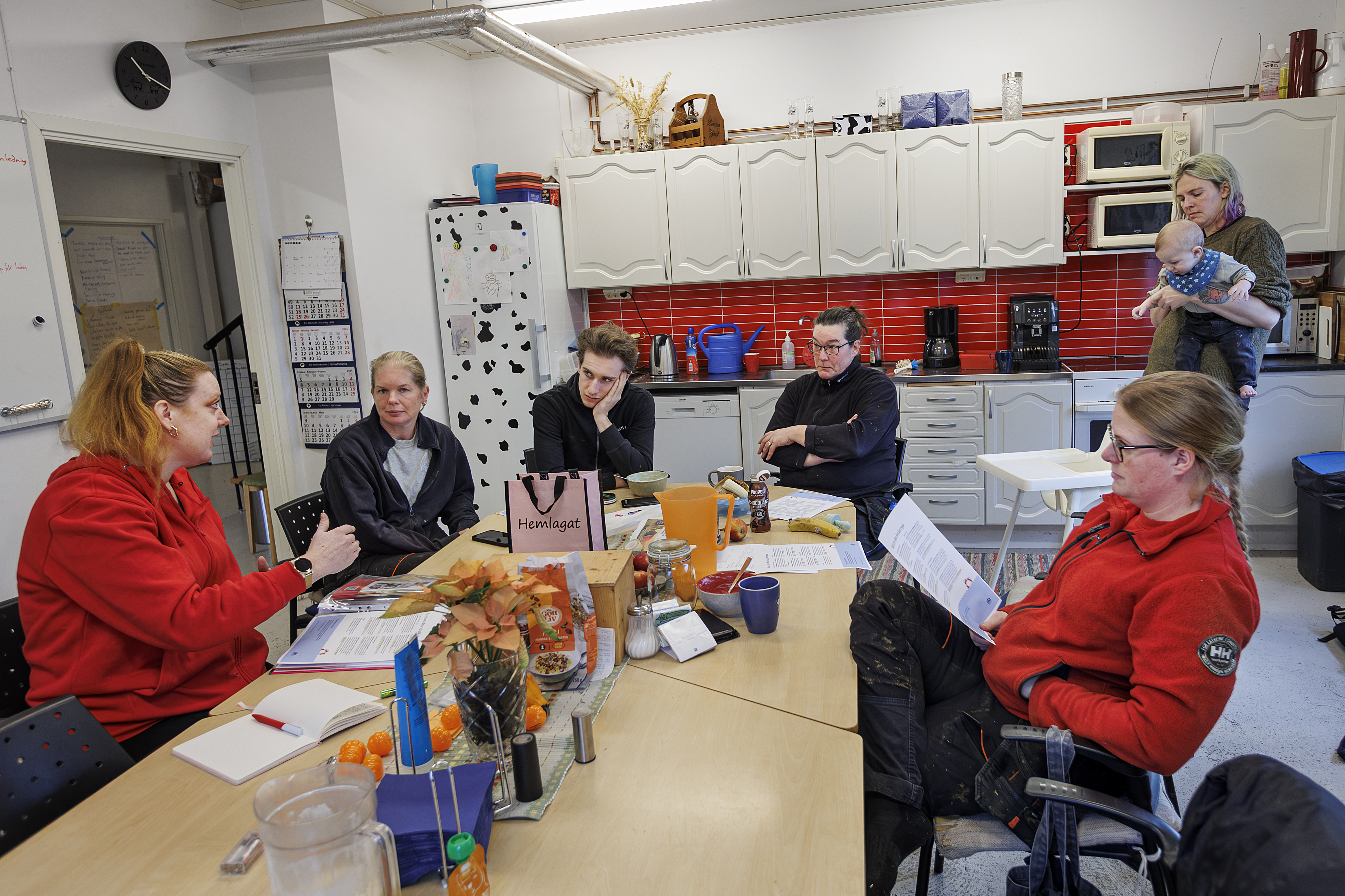 Sex vuxna sitter runt ett bord i ett kök och pratar; en person står i bakgrunden med en bebis. Papper och snacks ligger på bordet.