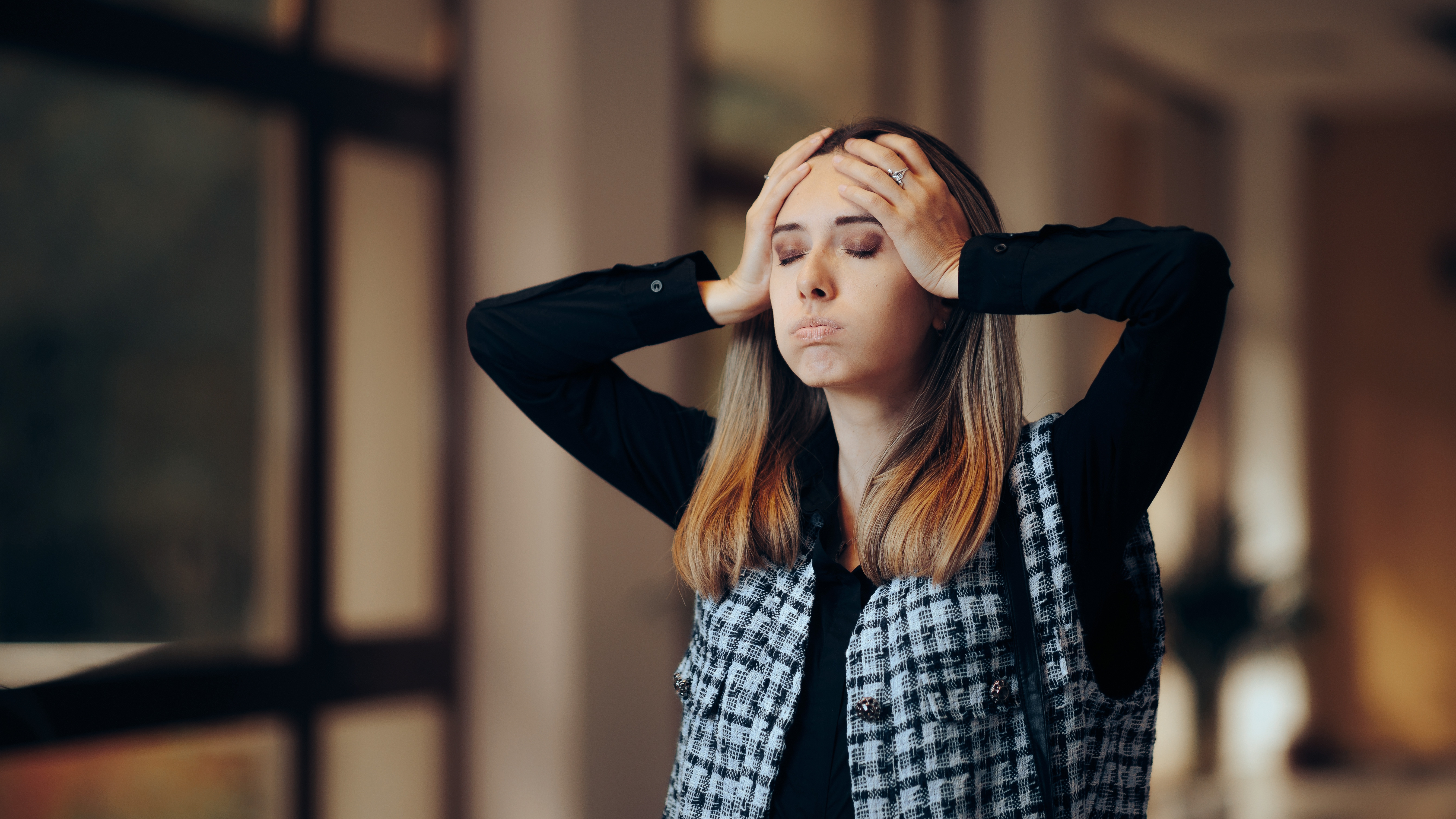 En kvinna står inomhus med händerna på huvudet och ögonen stängda.