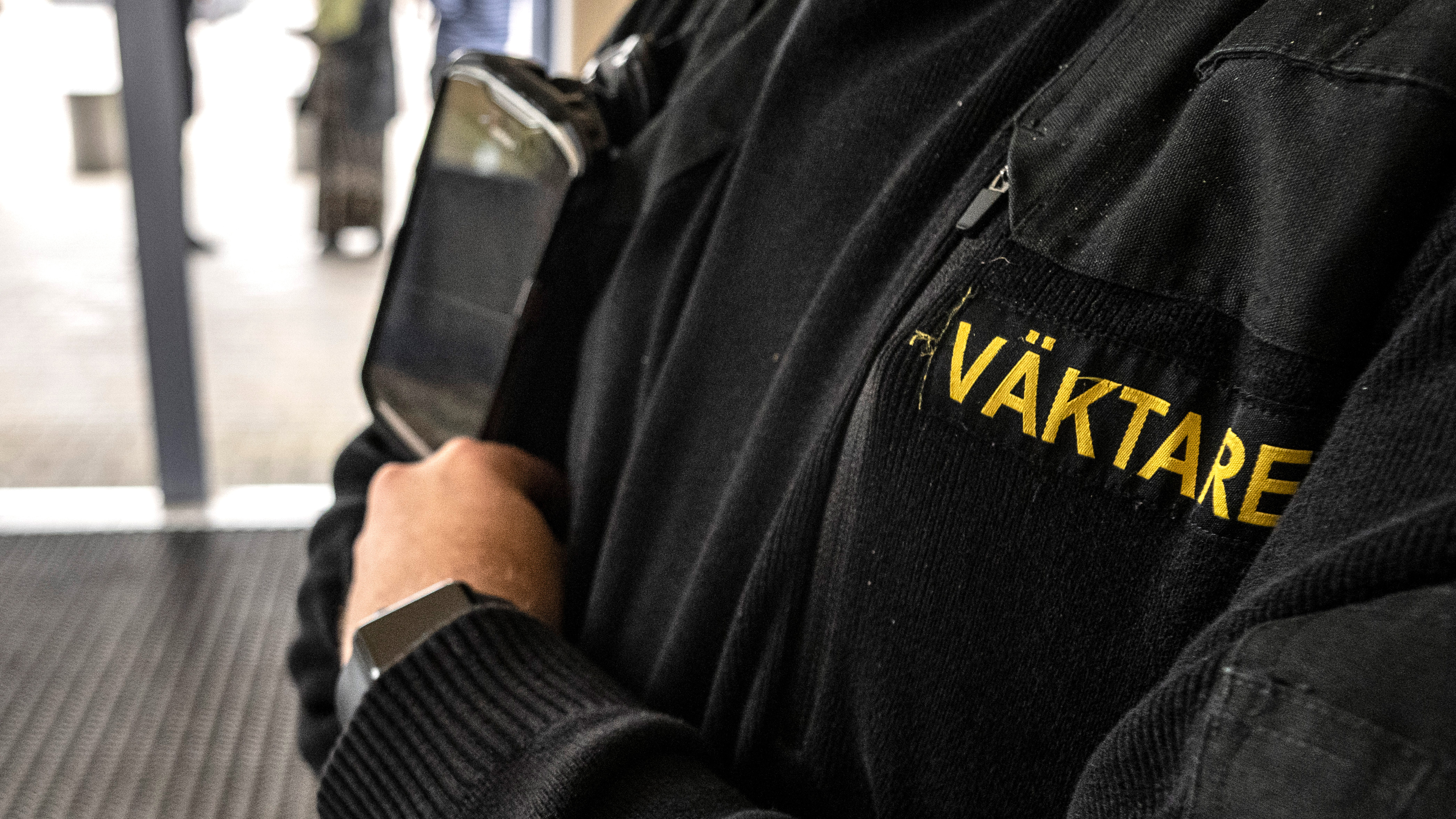 En person som bär en svart jacka som säger "väktare" Han har gula kläder på sig och håller en surfplatta eller enhet mot bröstet.