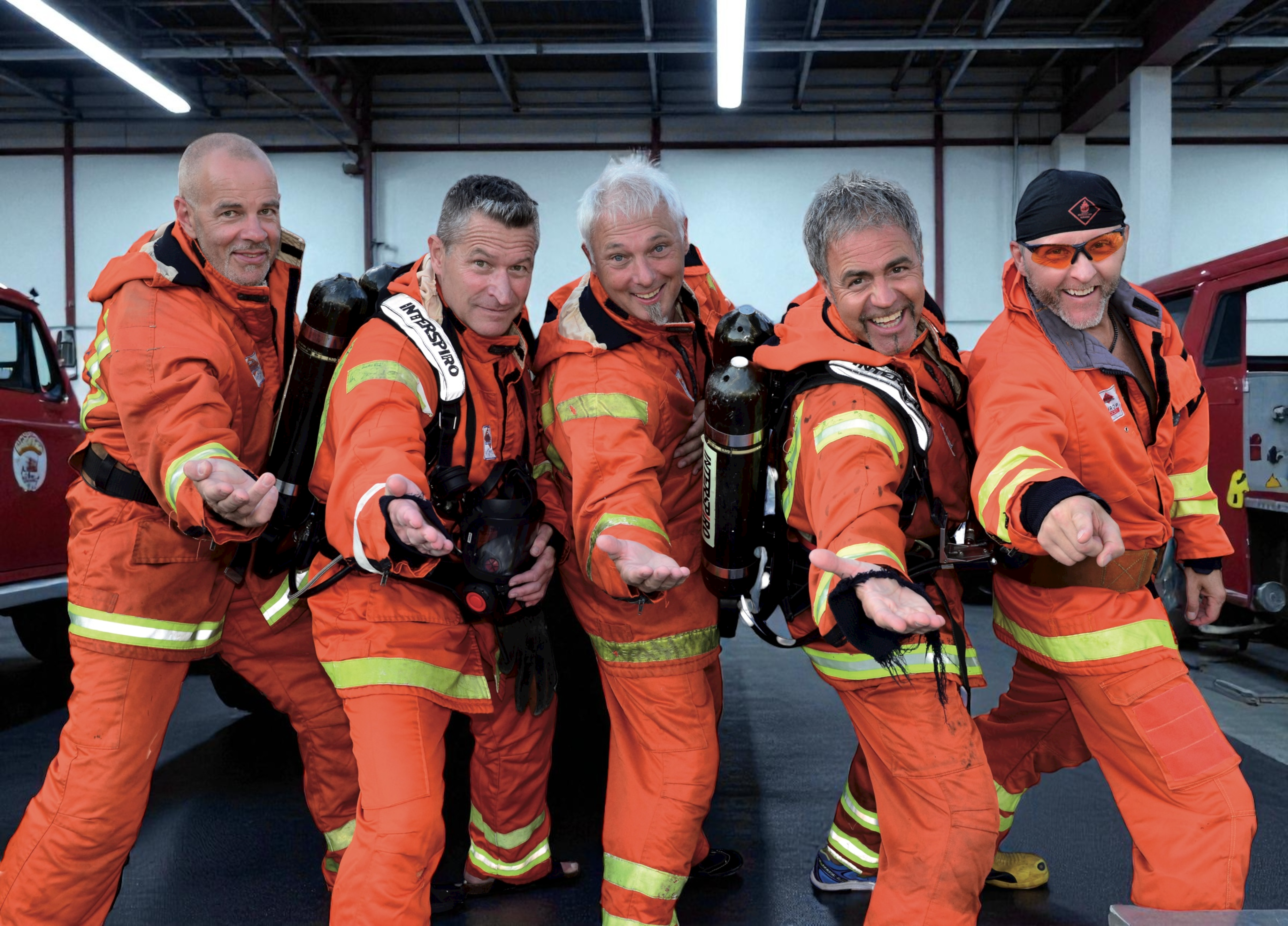 Fem män i orange brandmansuniformer, i bandet Brandsta city släckers, står tillsammans inomhus och sträcker fram en arm.
