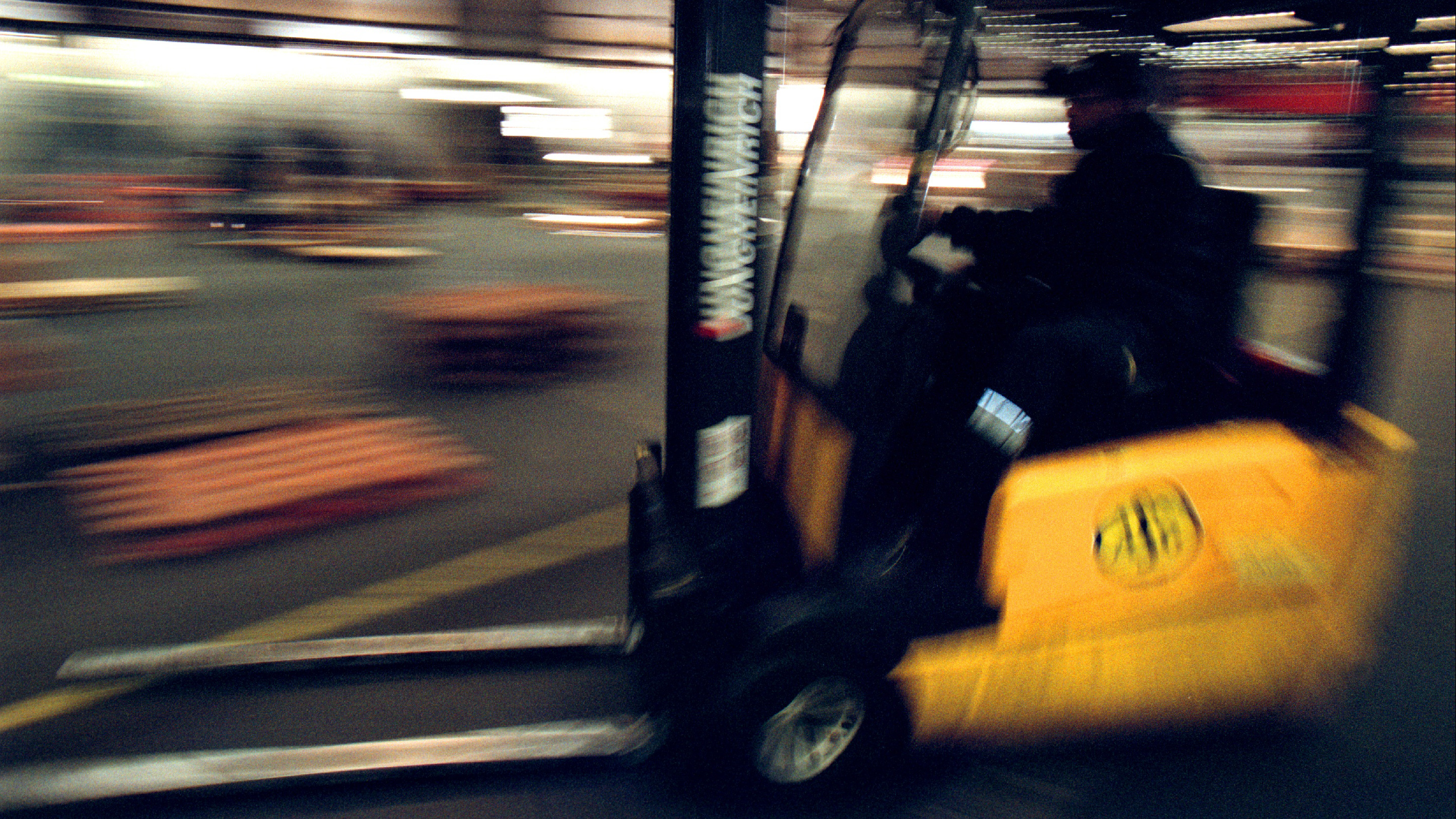 En person kör en gul gaffeltruck i ett lager. Personen är i rörelse och omgivningen är suddig.