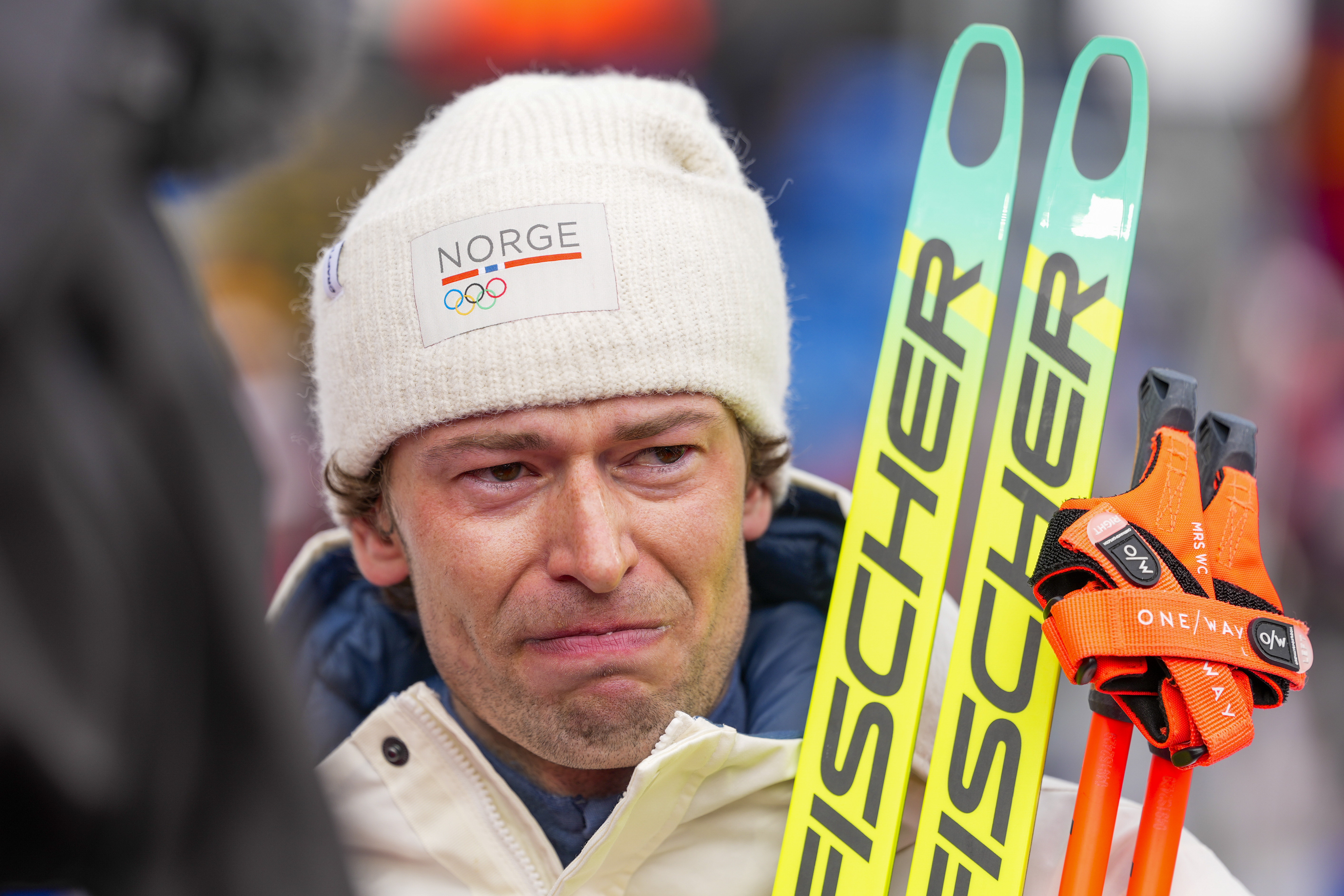 En skidåkare med en vit mössa med texten "Norge" håller Fischer-skidor vid ett utomhusevenemang och ser känslosam ut.