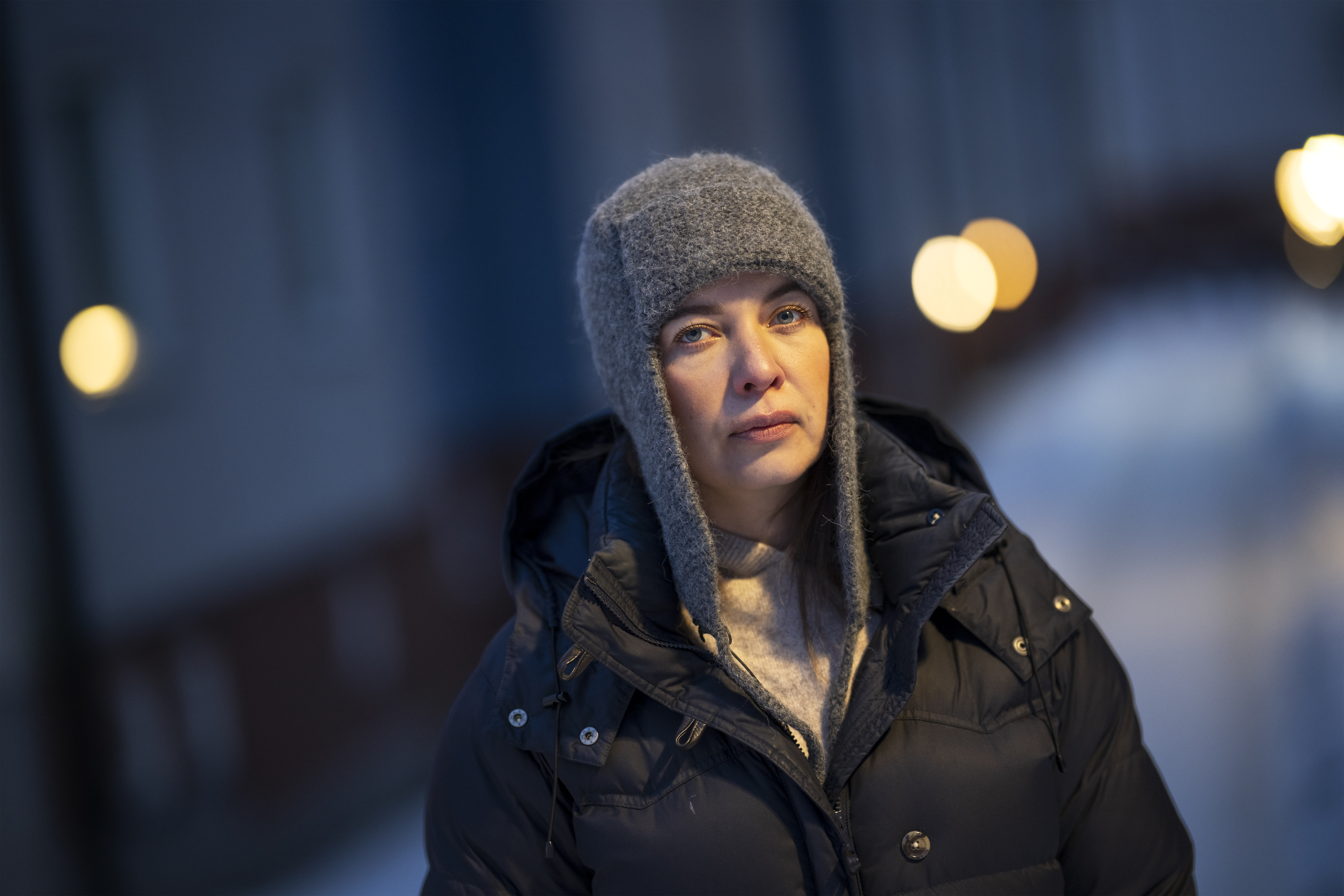 Lana, iklädd grå vintermössa och svart jacka, står utomhus i snön på kvällen eller natten, med ett suddigt ljus bakom sig.