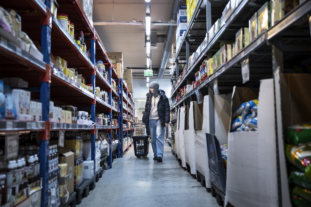 Lana, iklädd hatt och jacka, drar en kundvagn på hjul genom butiken mellan hyllorna.