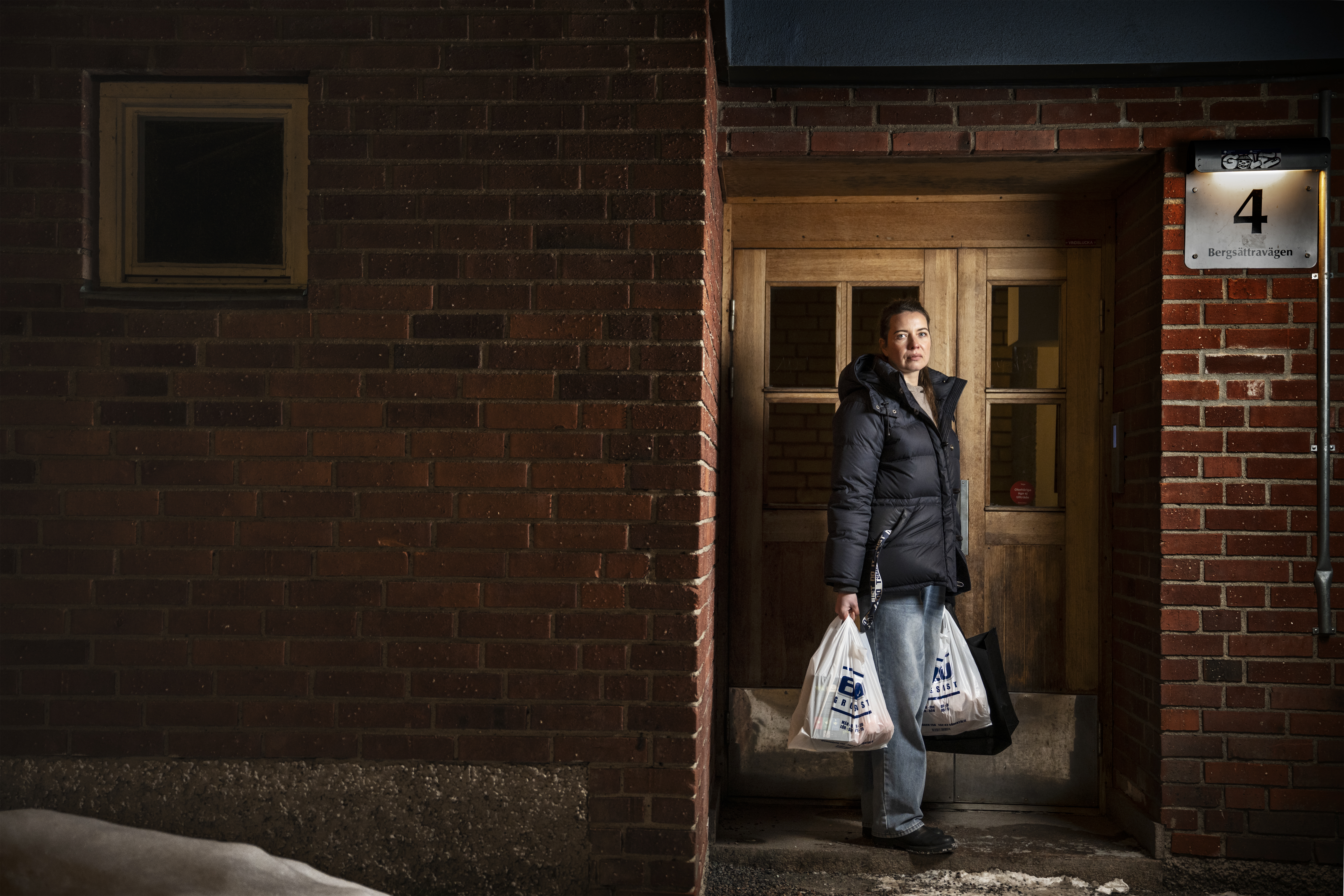 Lana, iklädd en svart rock, står i dörröppningen med två plastpåsar, utanför mot en tegelvägg och snö.