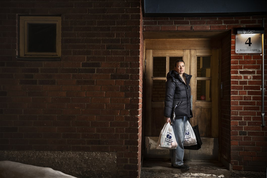 Lana, iklädd en svart rock, står i dörröppningen med två plastpåsar, utanför mot en tegelvägg och snö.