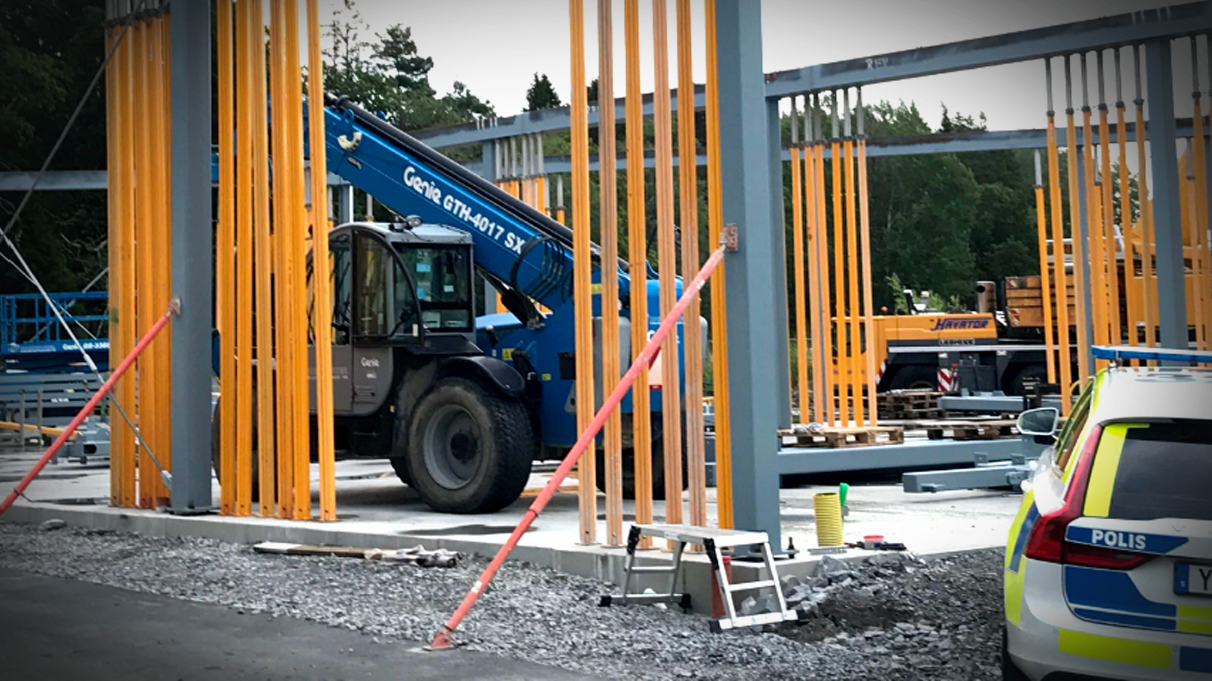 Byggarbetsplats med stålbalkar som installeras, en Genie teleskoplastare, stegar och en svensk polisbil i förgrunden.