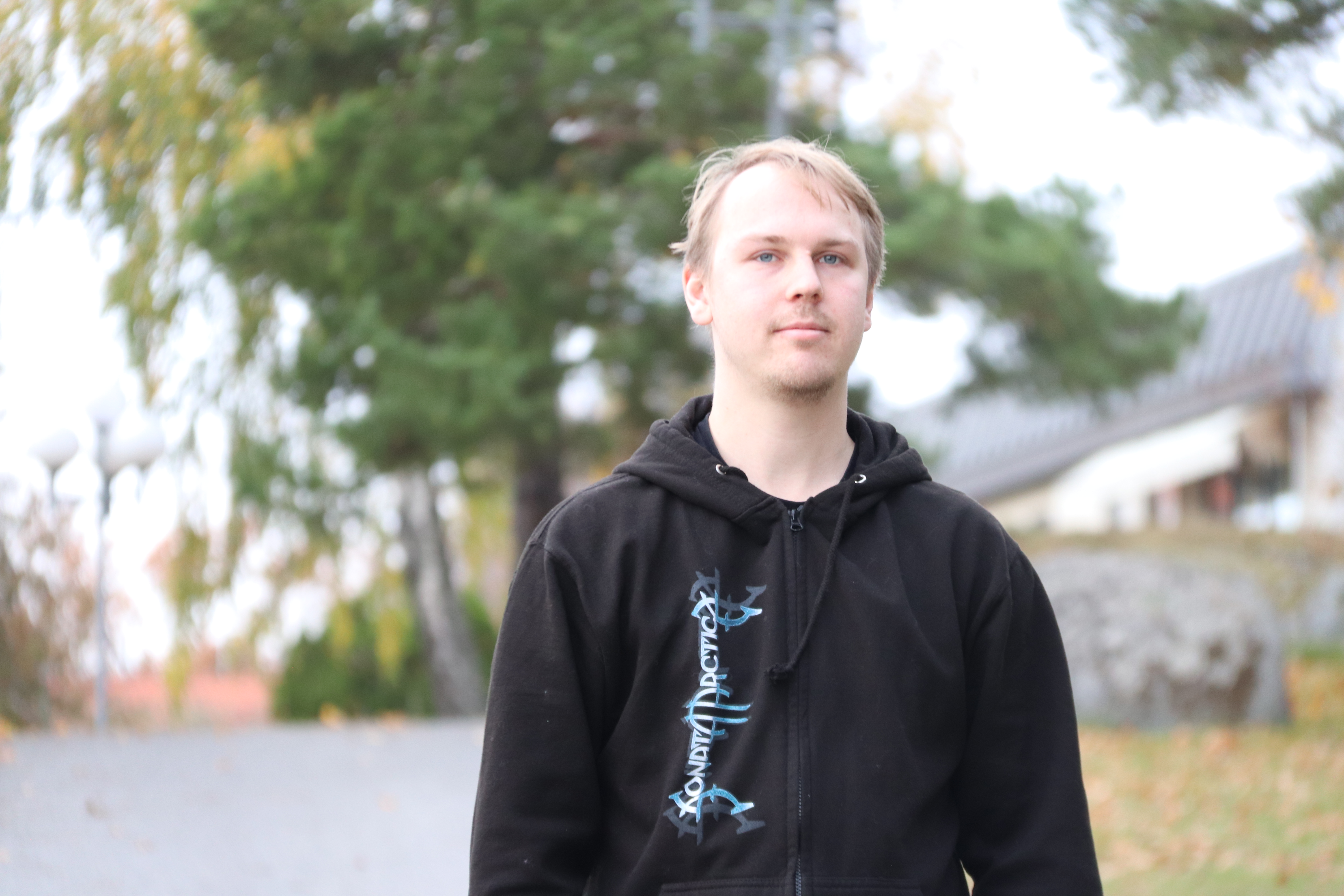 En ung man med blont hår och svart huvtröja står utomhus på en gångväg med träd och byggnader i bakgrunden.