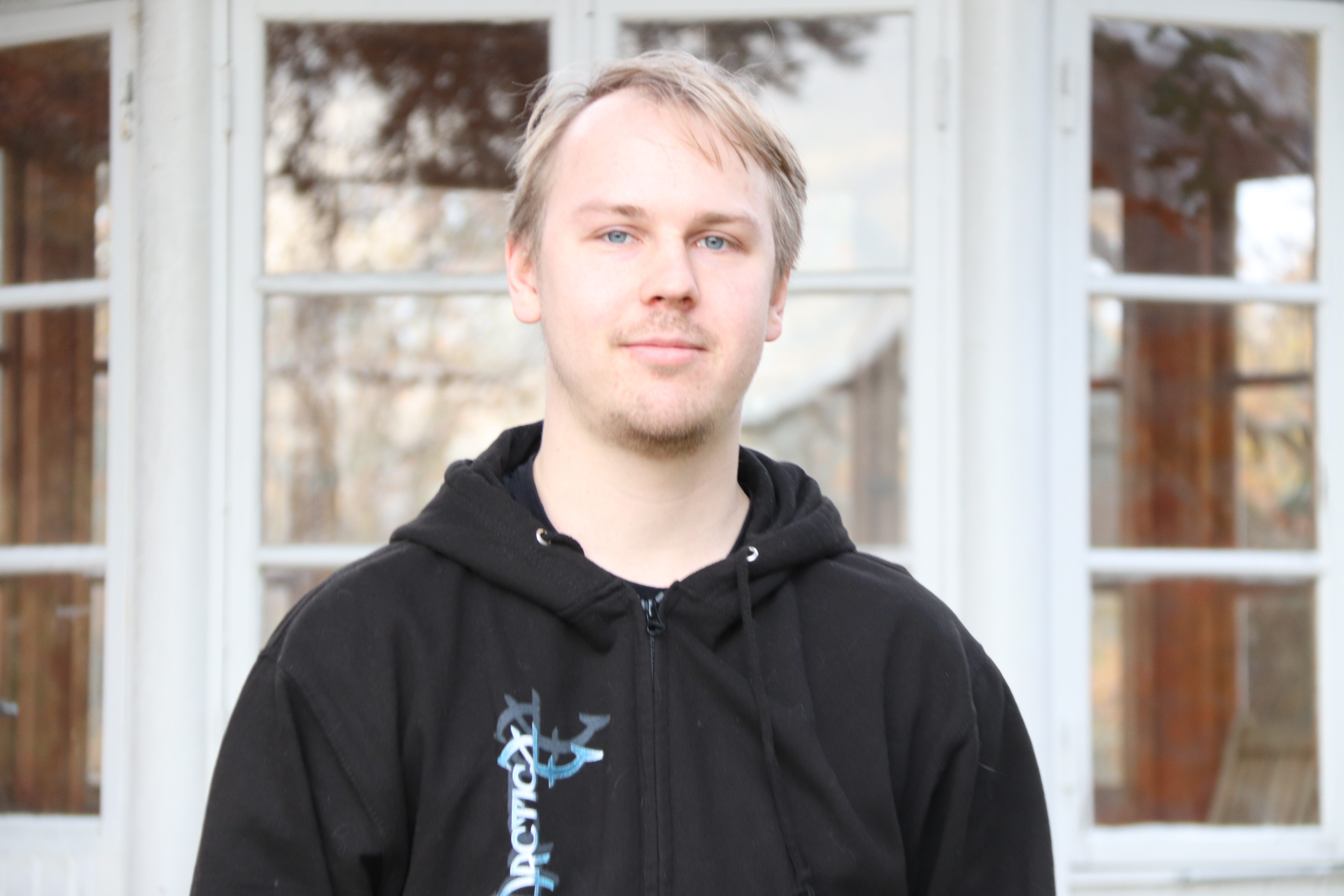 En ung man med kort, ljust hår har på sig en svart hoodie och står framför ett fönster med vit ram.