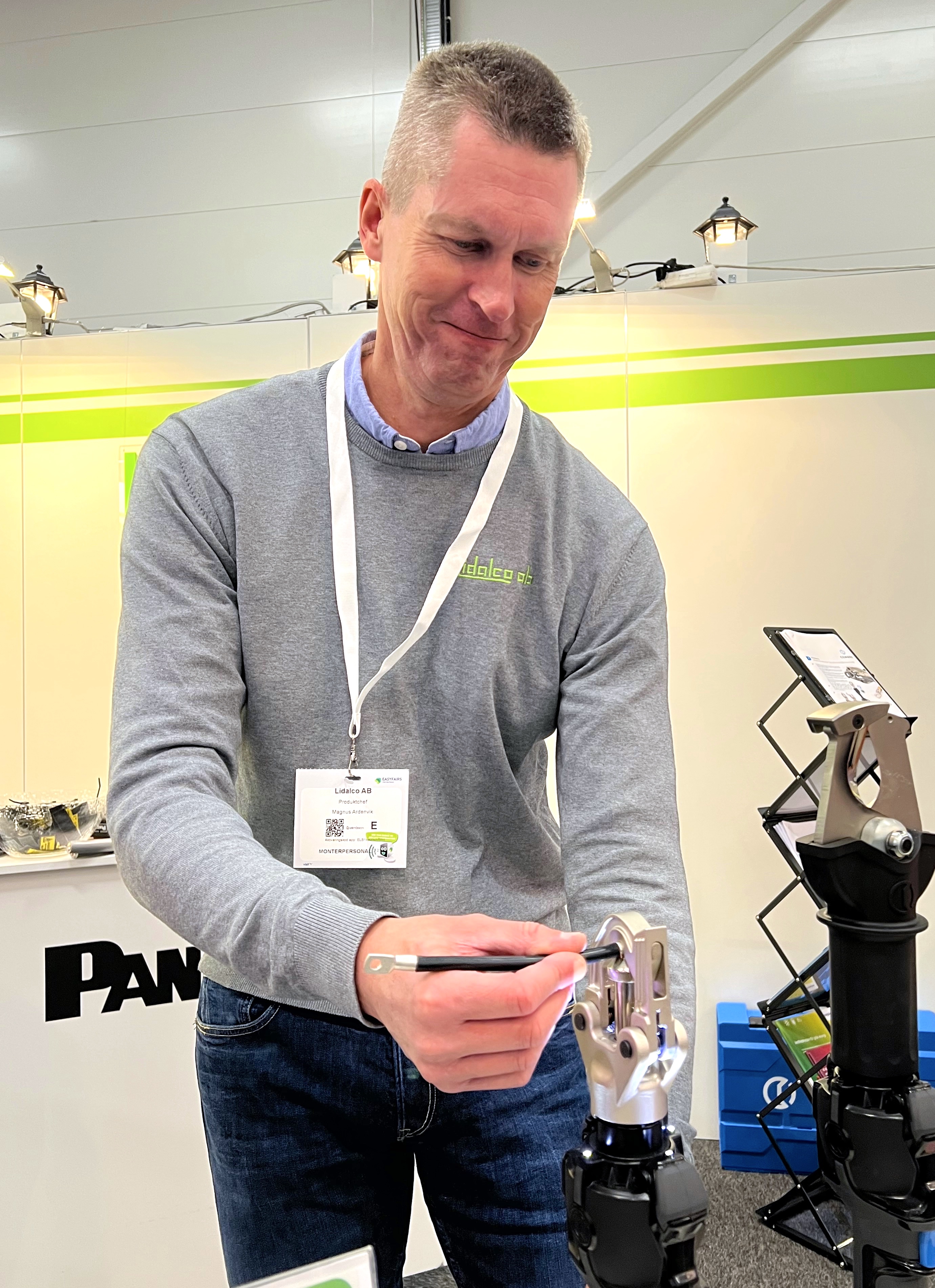 En man i grå tröja visar en mekanisk anordning med ett verktyg vid en monter, bär konferensbricka, bakgrunden har ställ och utrustning.