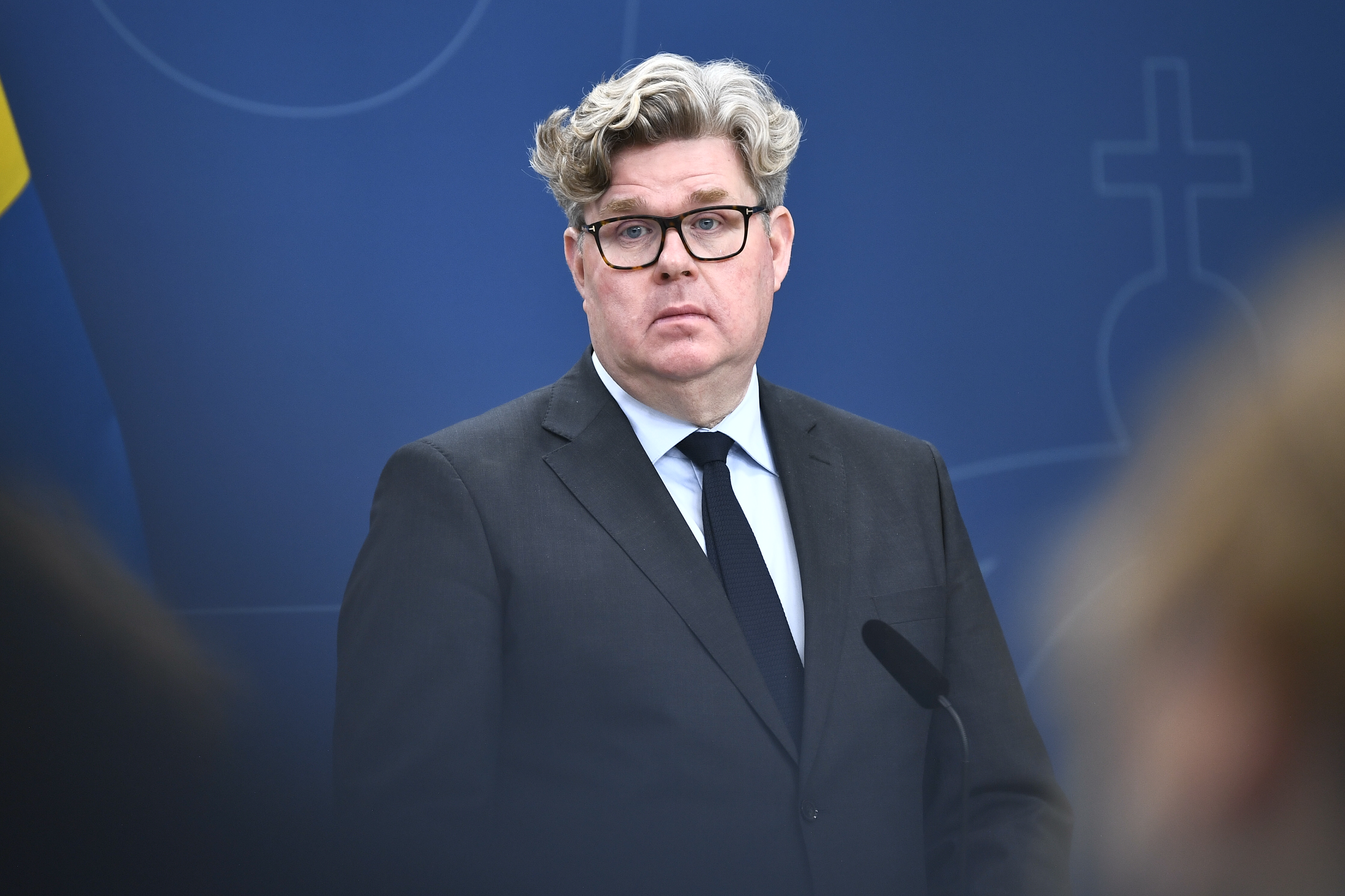 En man med glasögon och mörk kostym står framför en blå bakgrund med en mikrofon.