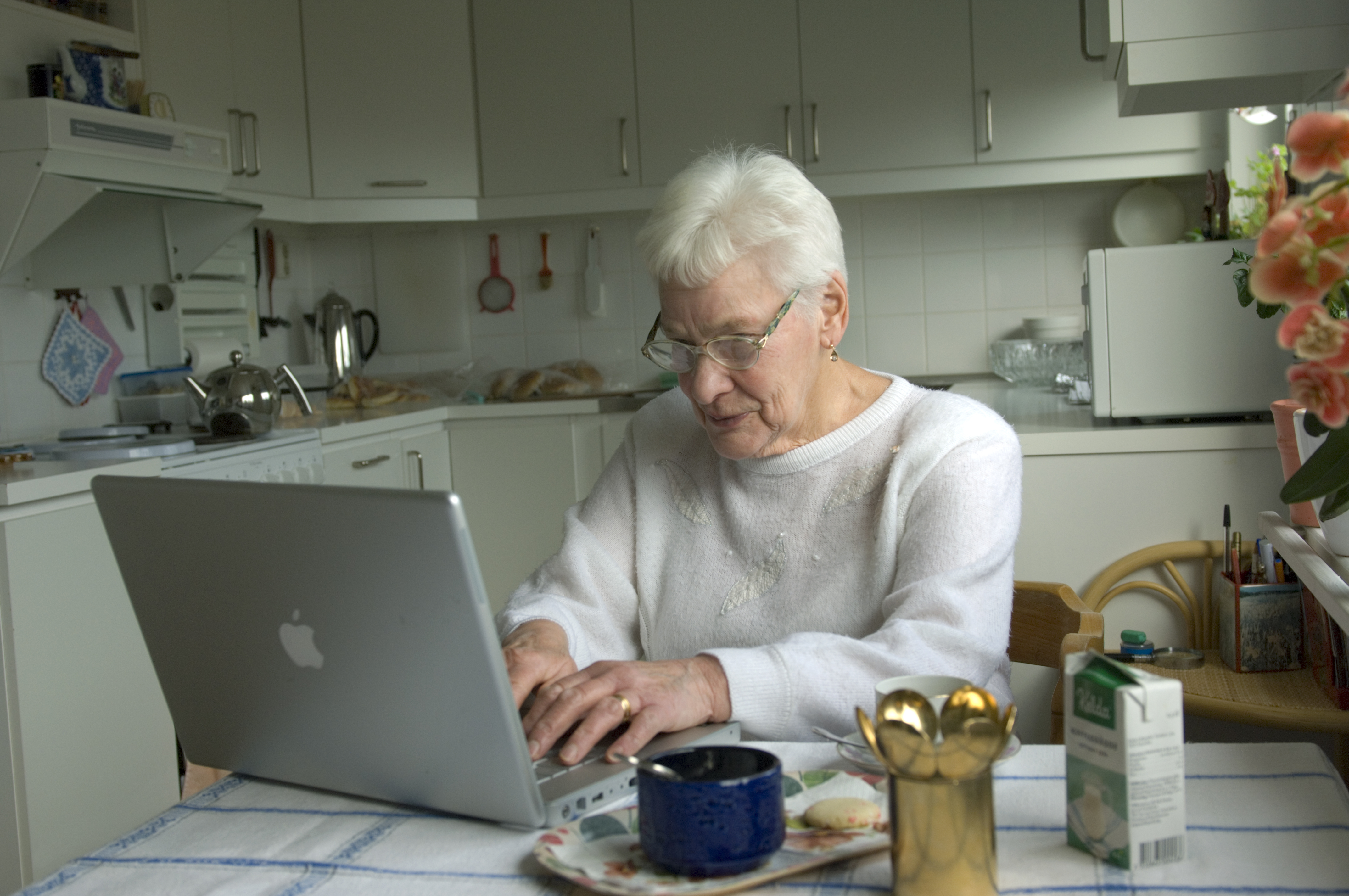 En äldre kvinna sitter vid ett köksbord och använder en bärbar dator. Bredvid henne finns te och tilltugg i köket.