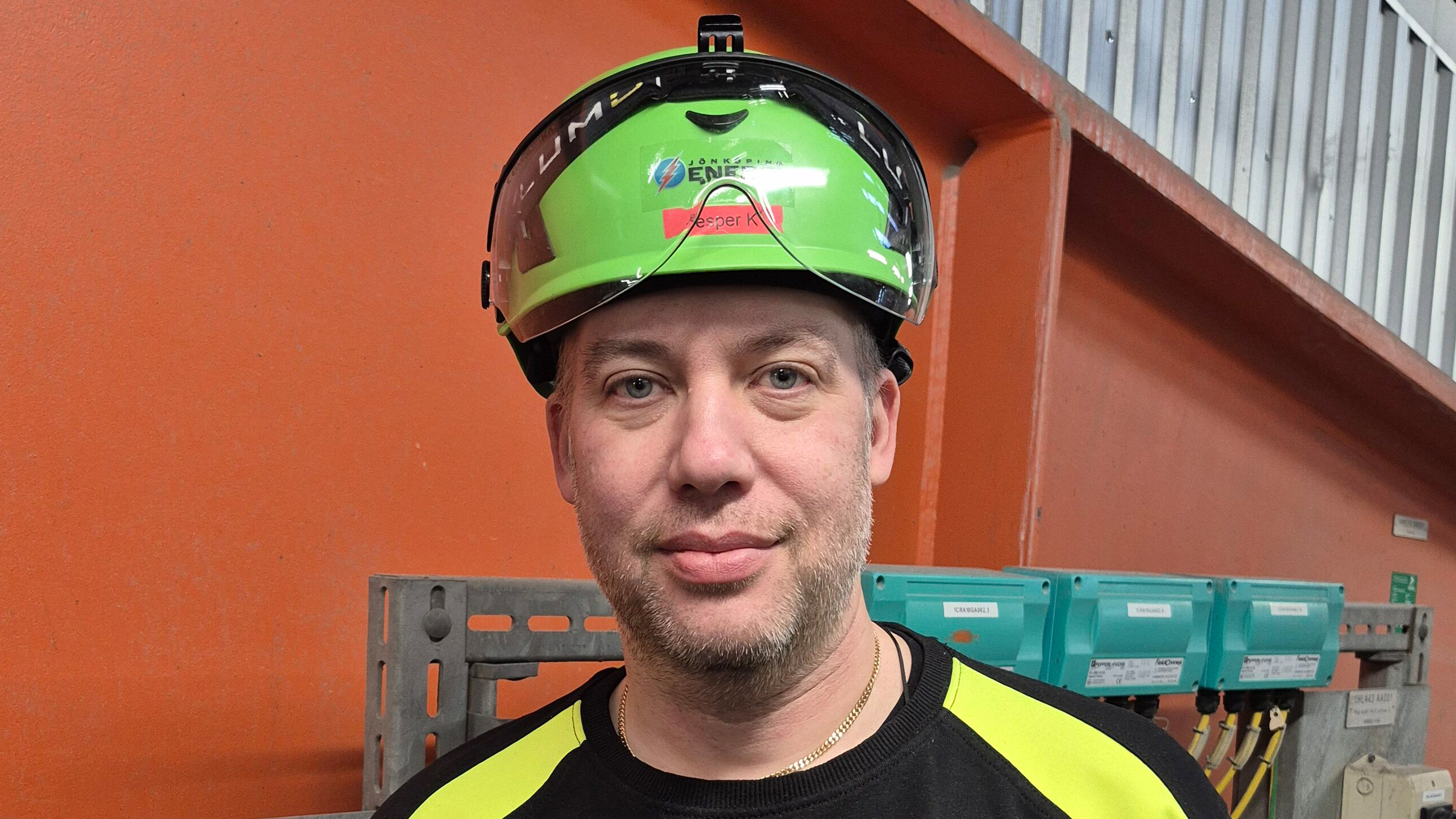 Man med grönt skyddshjälm och varselkläder står inomhus nära orangea och metallväggar med elutrustning i bakgrunden.