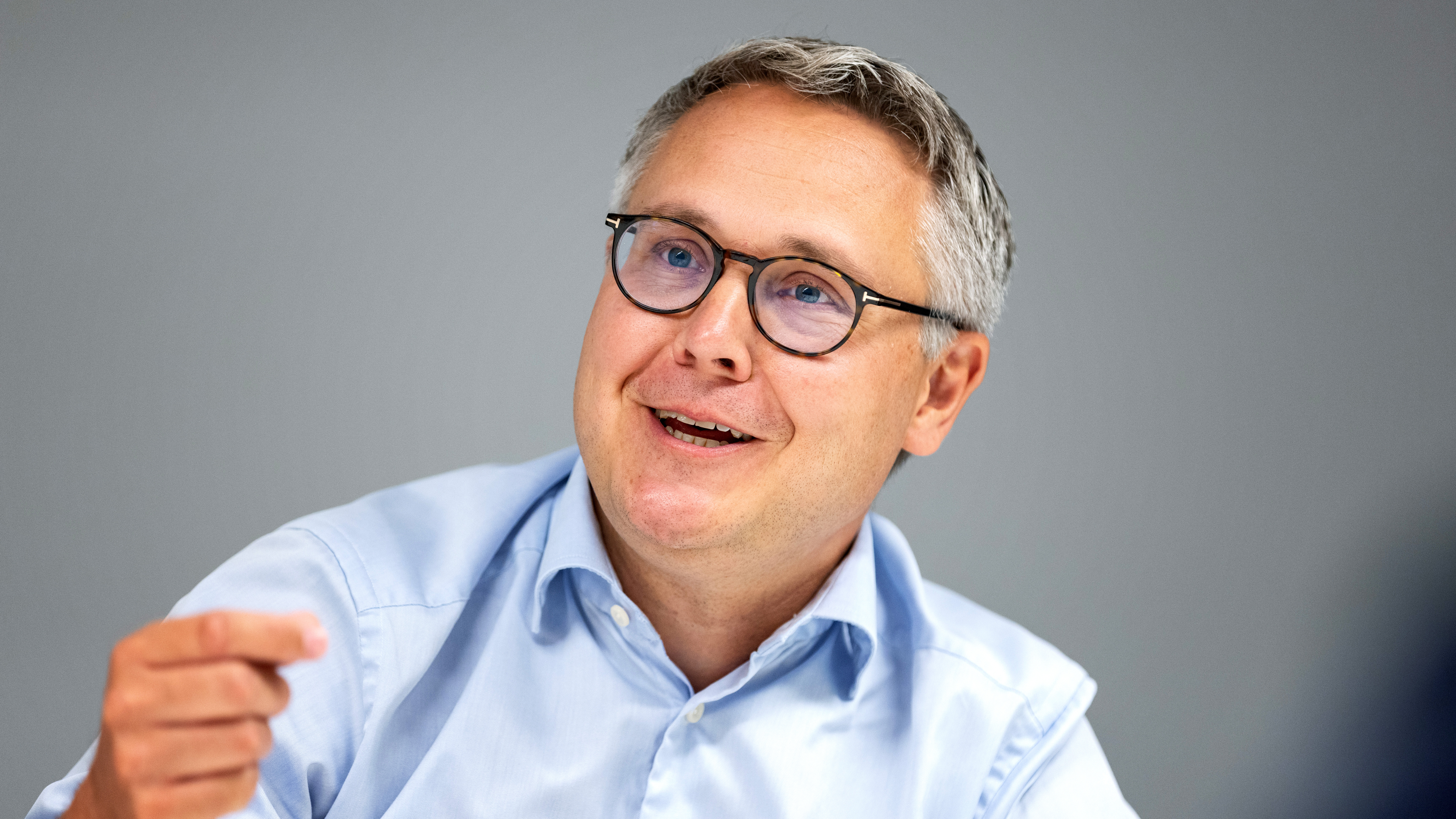 Johan Danielsson med kort grått hår och glasögon, iklädd en ljusblå skjorta, ler och gör en gest med handen mot en enkel bakgrund.