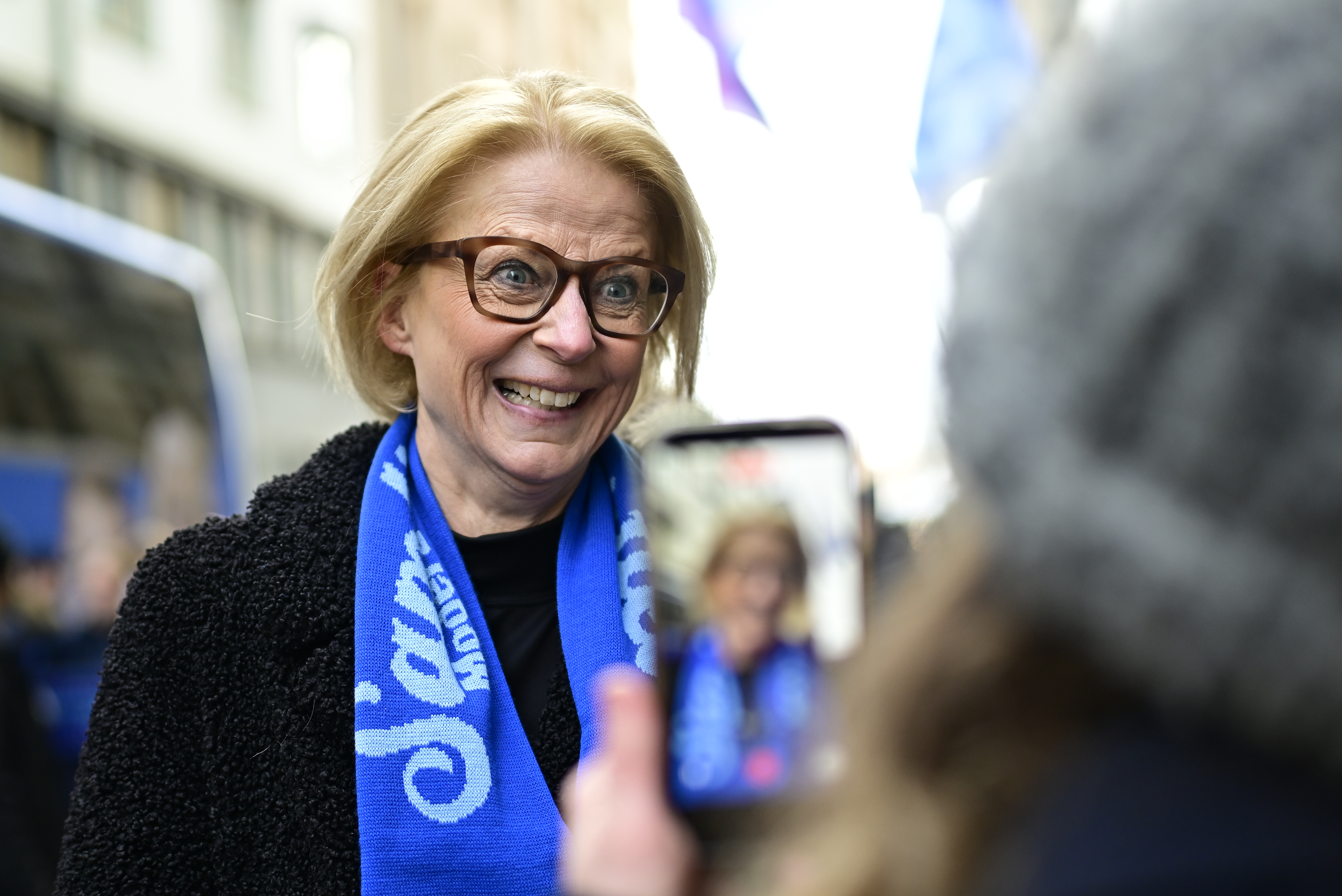 En kvinna med glasögon och blå scarf tittar på en person som fotograferar henne med en mobil utomhus.