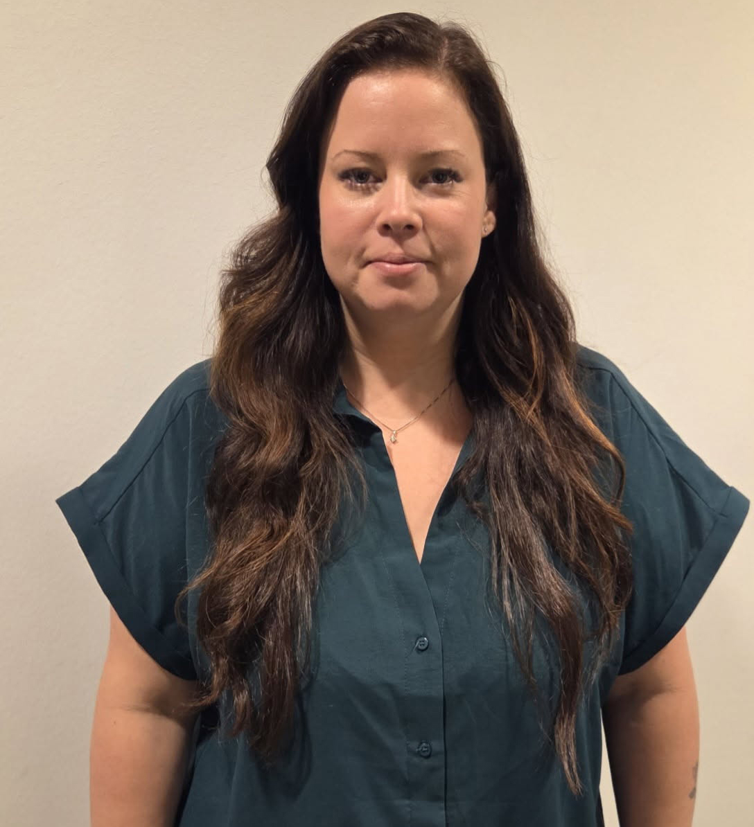 En kvinna med långt brunt hår och en mörkt tealfärgad skjorta står mot en ljus vägg och tittar mot kameran.