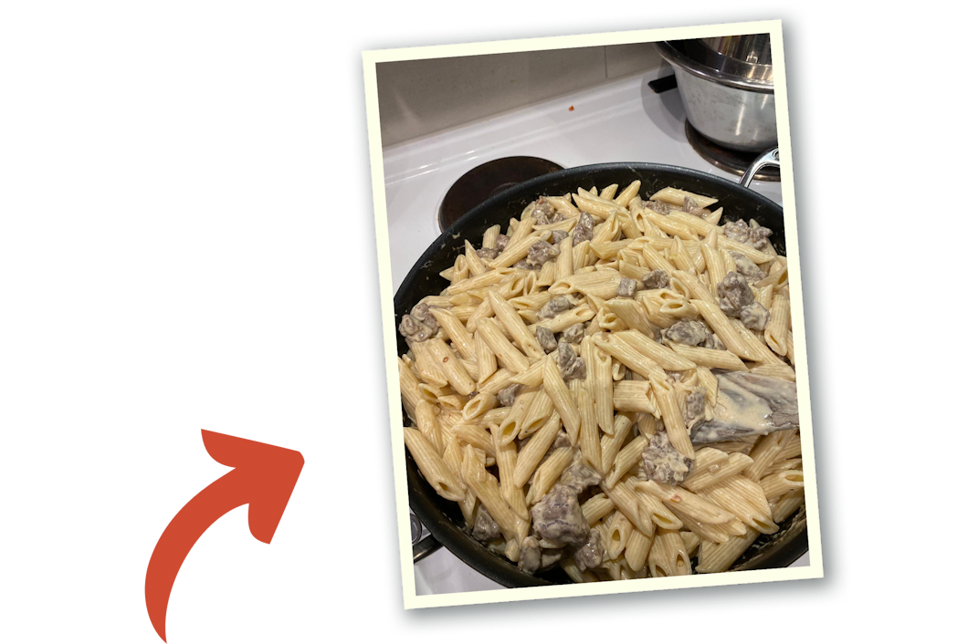 En stekpanna med pasta, sås och biffbitar står på spisen.