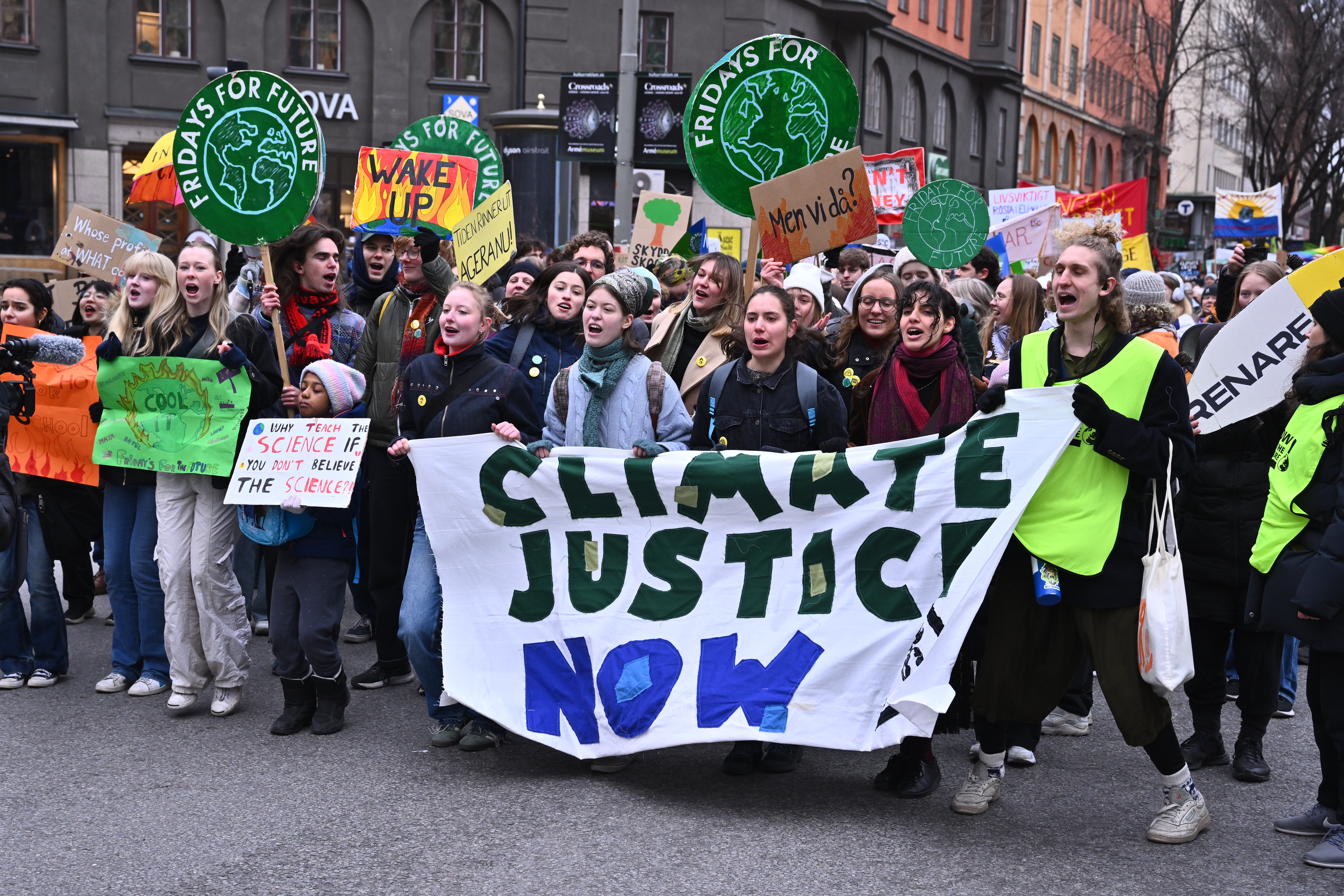 En grupp demonstranter marscherar med skyltar och en banderoll med texten "Climate Justice Now" under en klimatdemonstration i stadsmiljö.