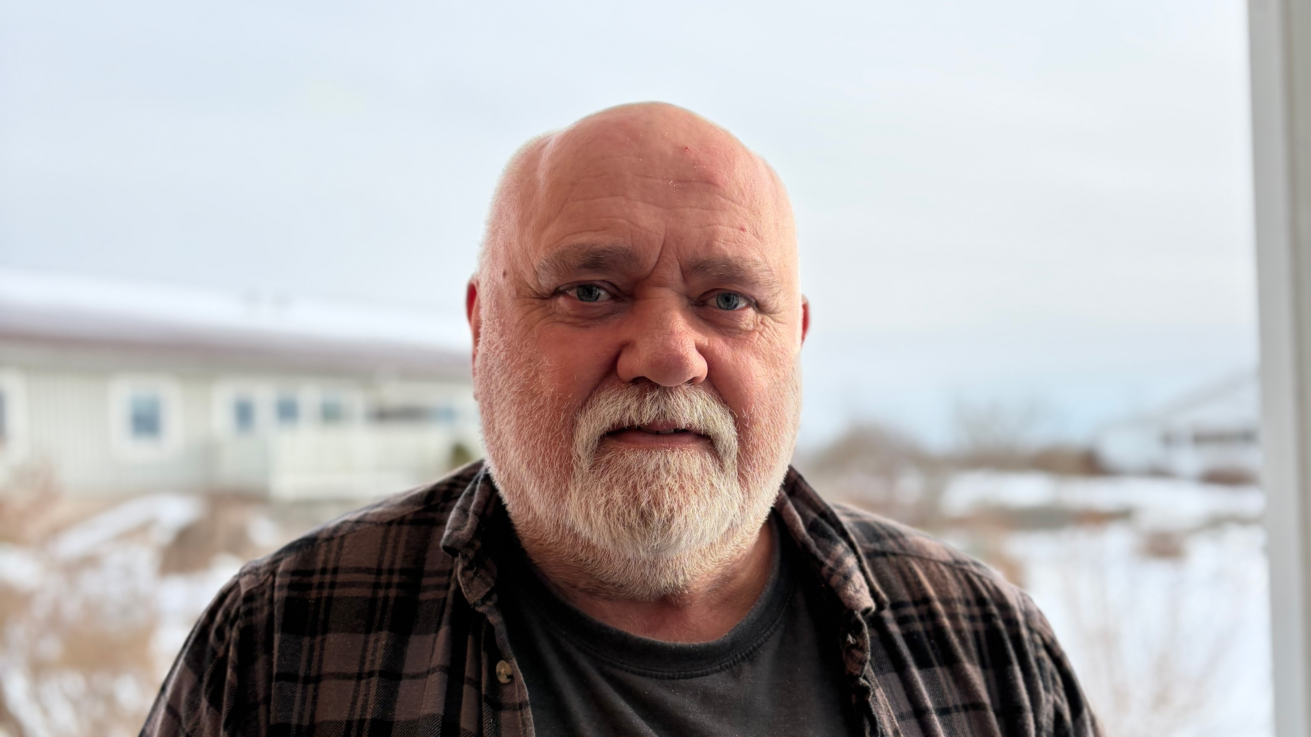 En gammal man med vitt skägg och flintskallig, iklädd rutig skjorta, står utomhus framför ett hus med ett snöigt landskap.