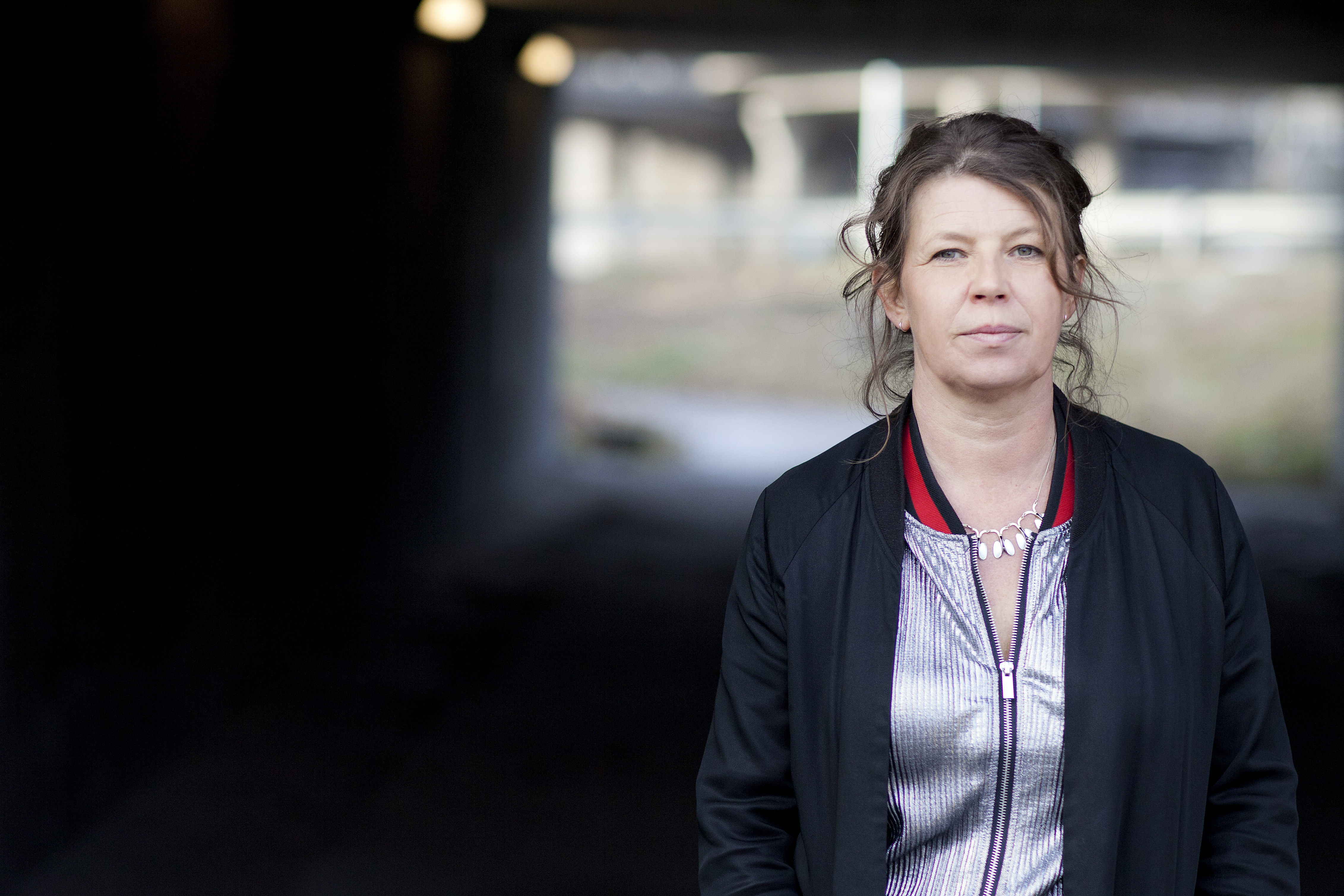 Poeten Lina Ekdahl, med brunt hår och svart jacka och randig skjorta står framför en tunnel med suddiga lampor och bakgrund.