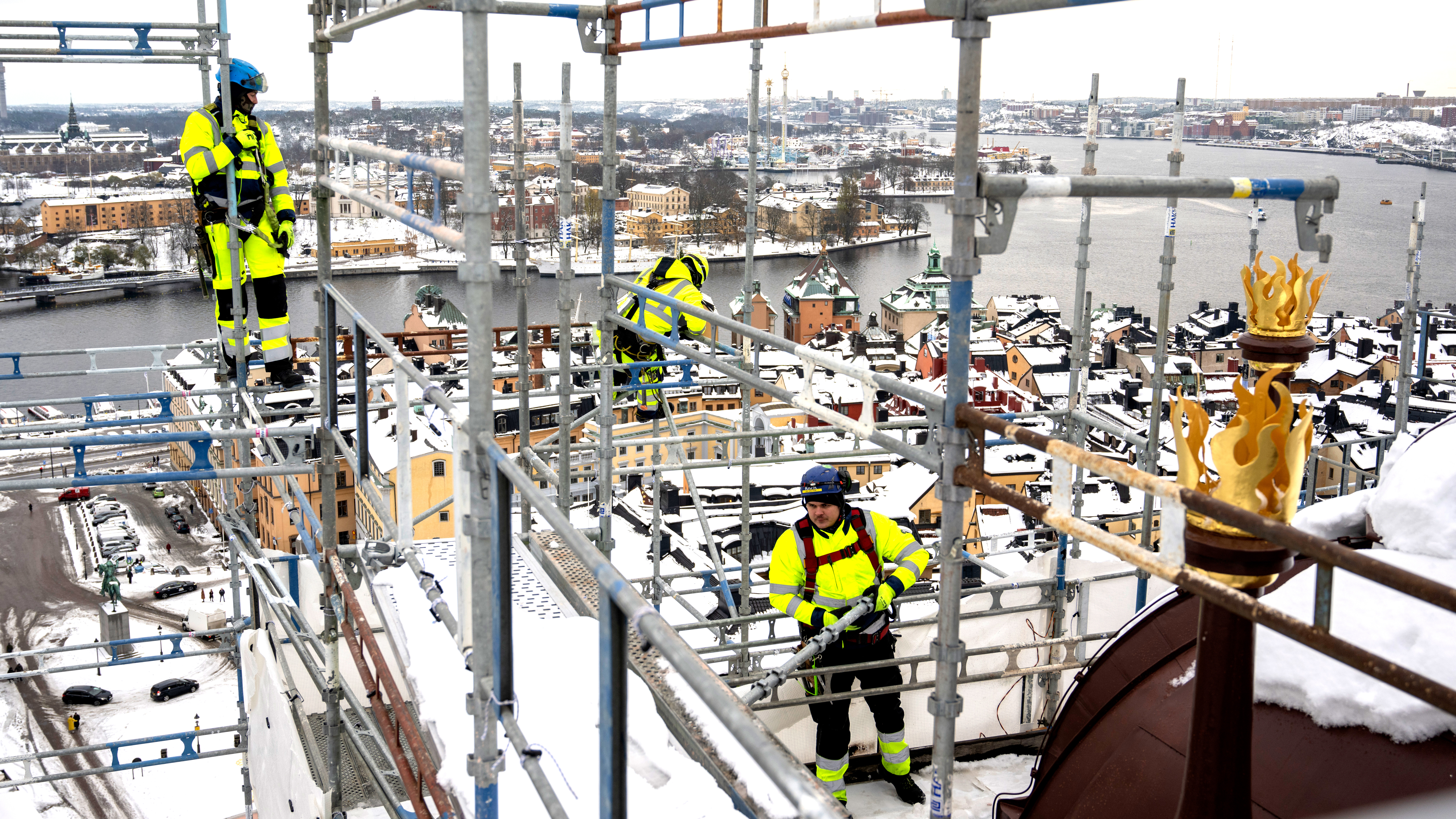 Tre byggarbetare i varselkläder står på en ställning över en snötäckt stad vid en flod, med byggnader under sig.