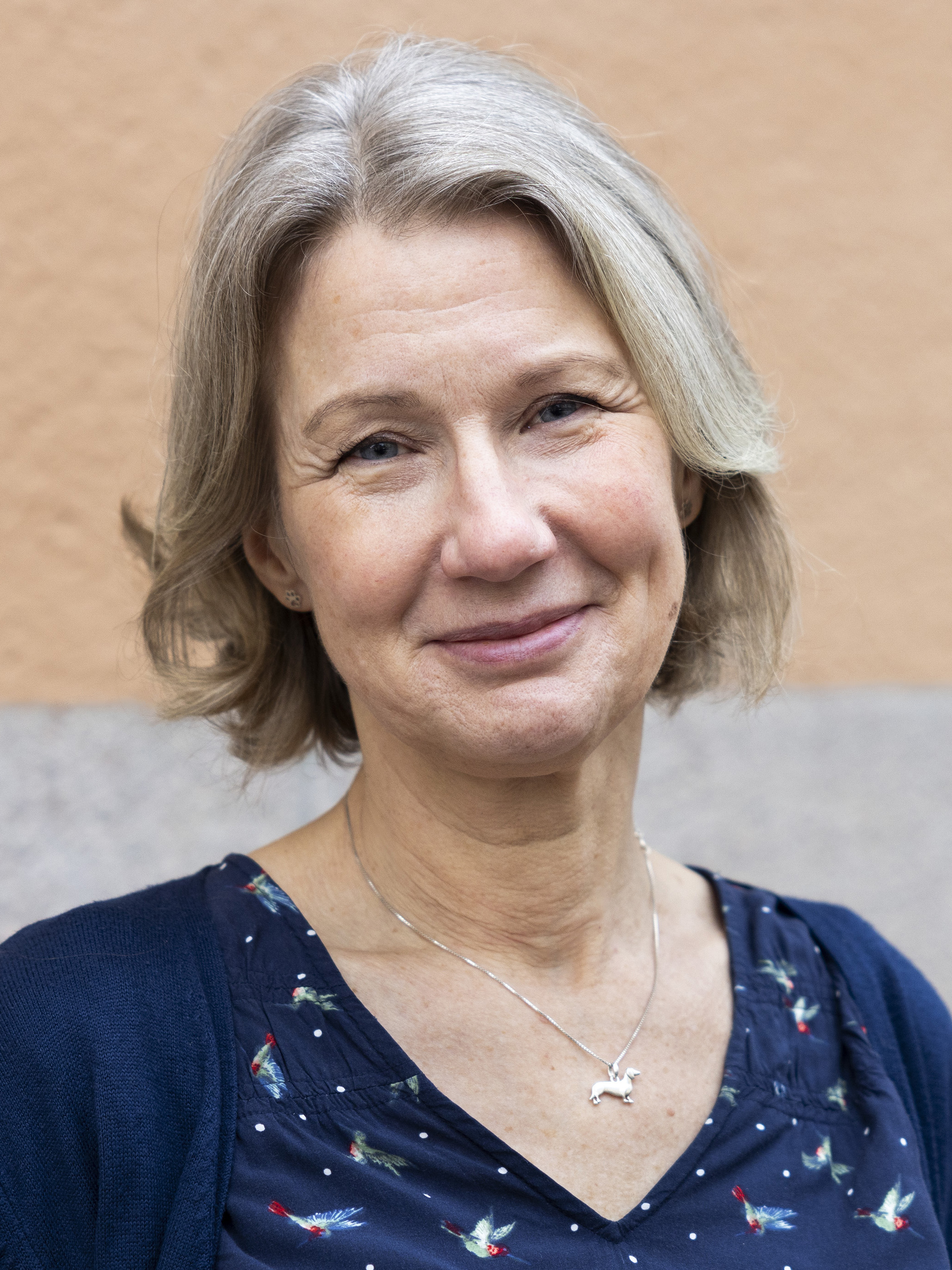 Carina Mood med kort grått hår ler, har en mönstrad blus, mörk kofta och silverhalsband, framför en vägg.