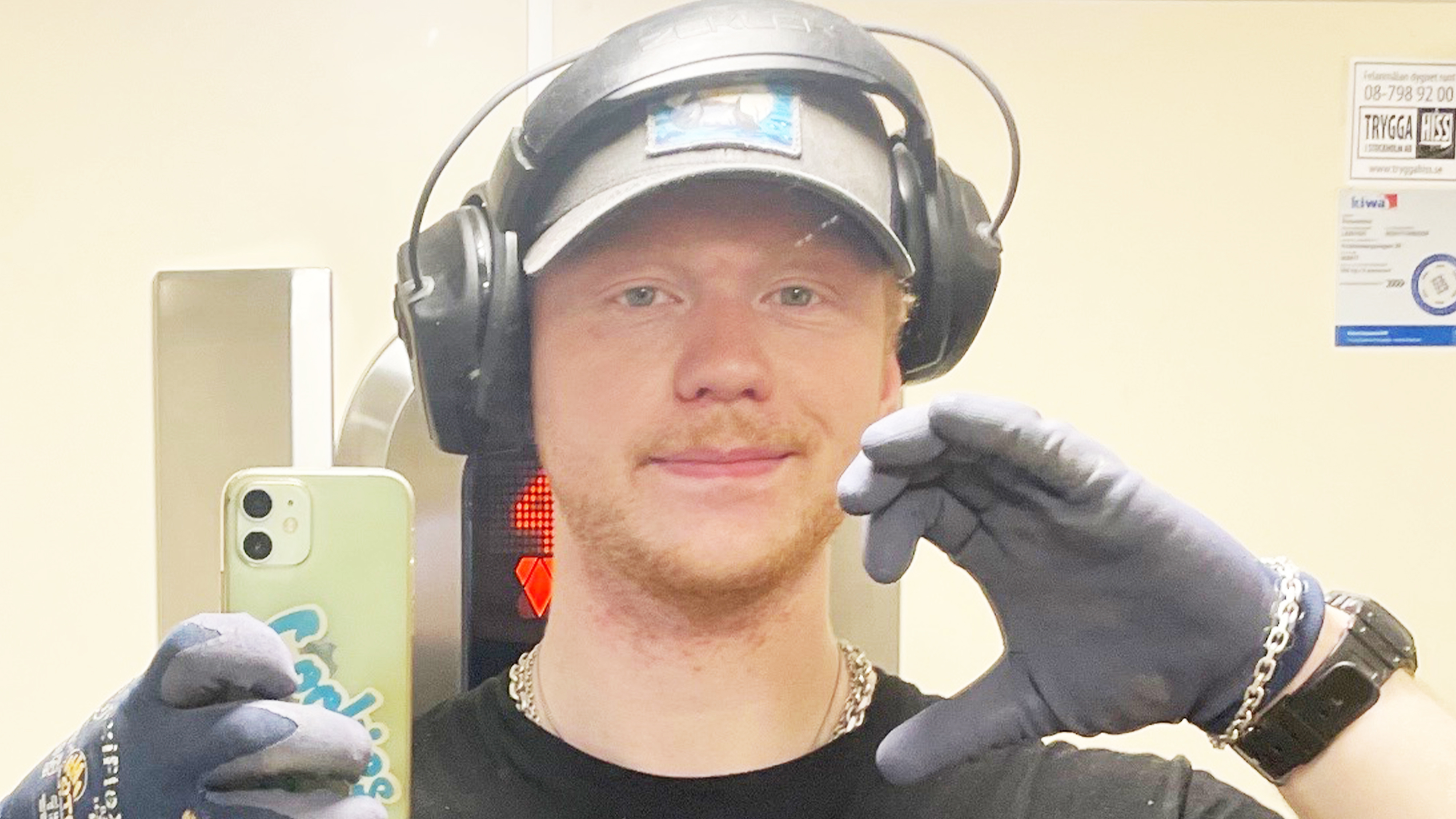 En person med handskar, hörlurar och kepsar tar en spegelselfie med en mobil och gör en handgest nära ansiktet.