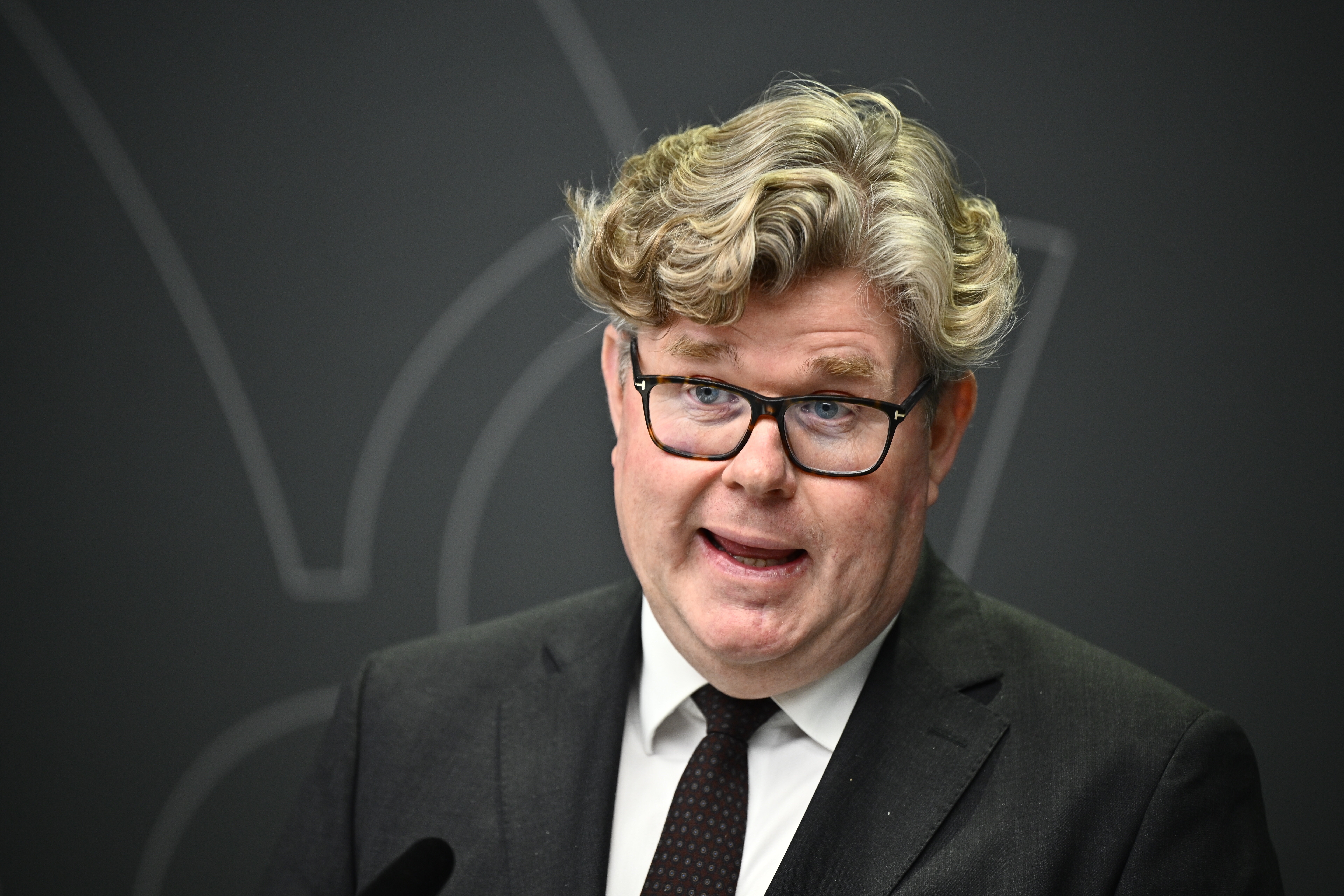 En man med lockigt hår, glasögon och kostym talar framför en mörk bakgrund med ett svagt abstrakt mönster.