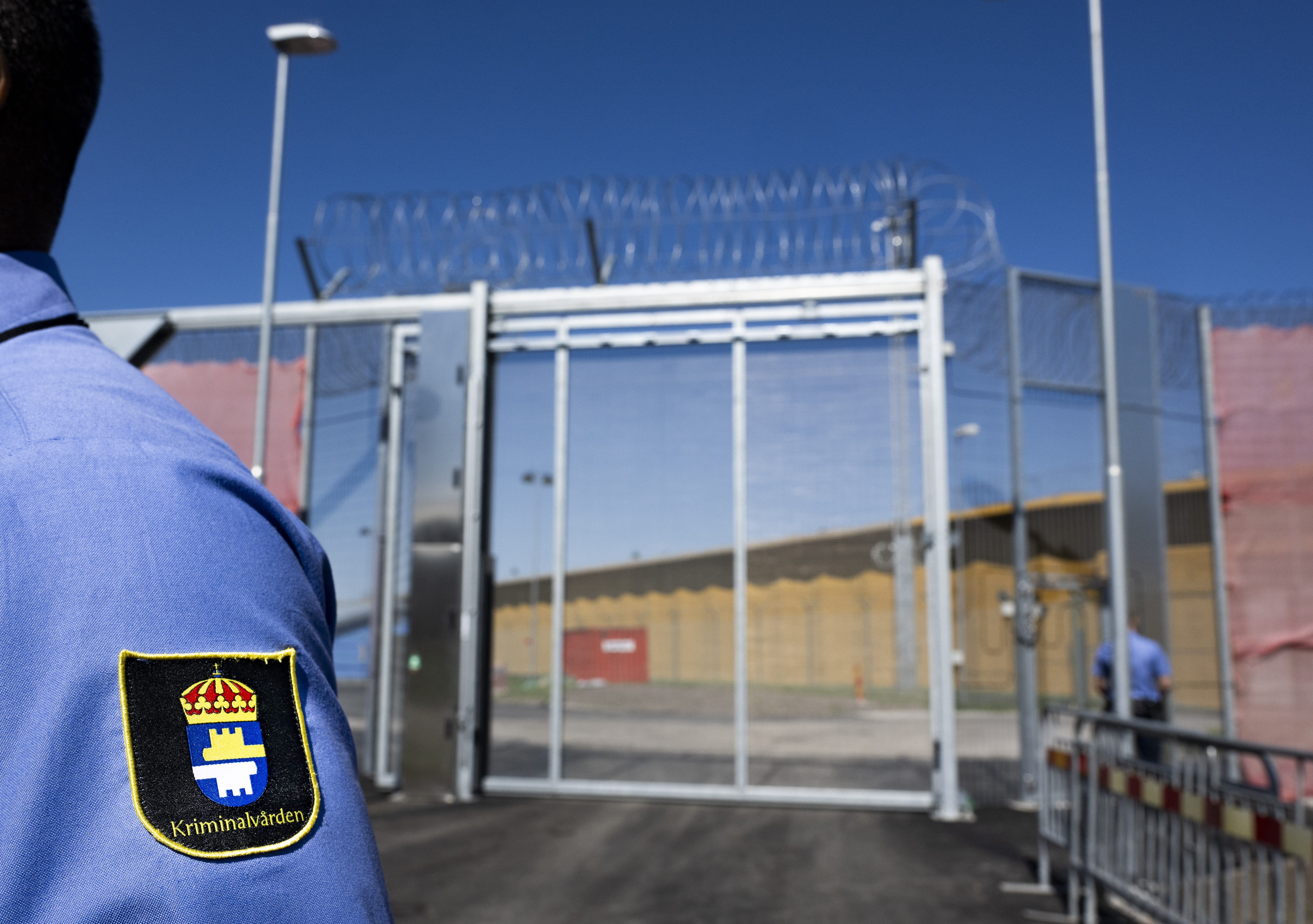 En säkerhetsvakt står framför ett stängsel med taggtråd och en grind vid en anläggning, med märket "Kriminalvården" på uniformen.