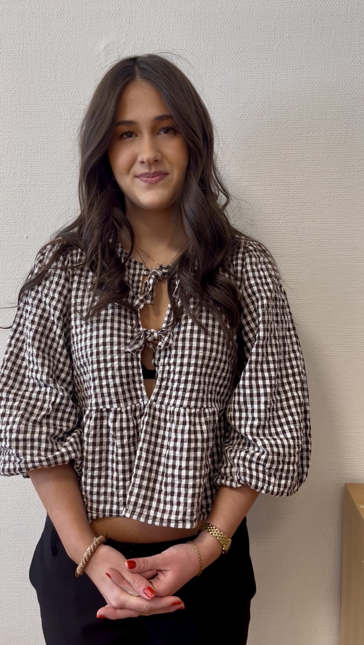 En kvinna med långt brunt hår står mot en vit vägg, har en brun- och vitrutigt blus med puffärmar och svarta byxor.