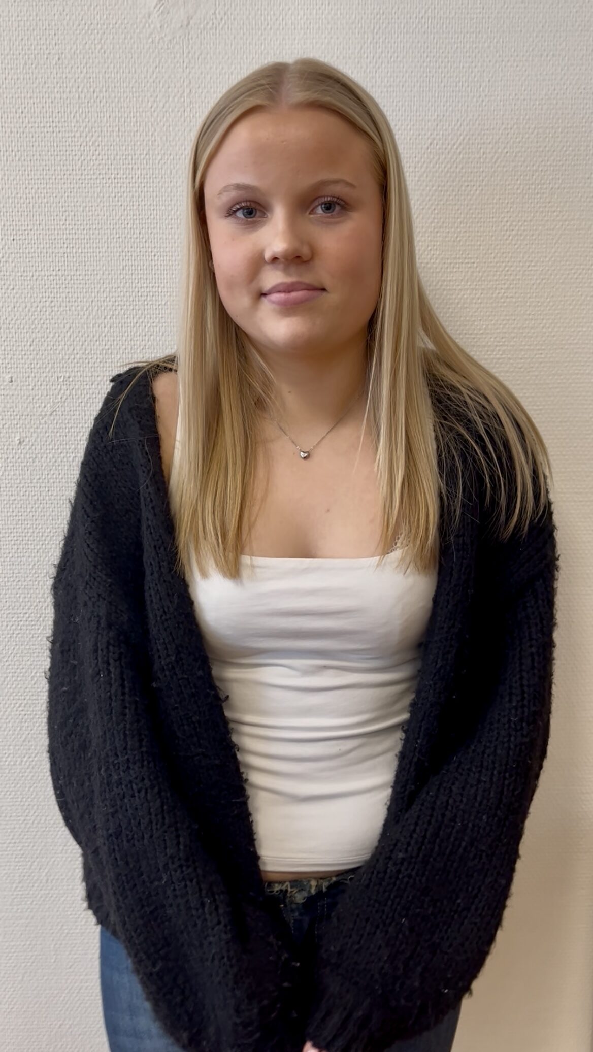 En ung kvinna med rakt blont hår, vit topp och svart kofta står mot en ljus vägg och tittar mot kameran.