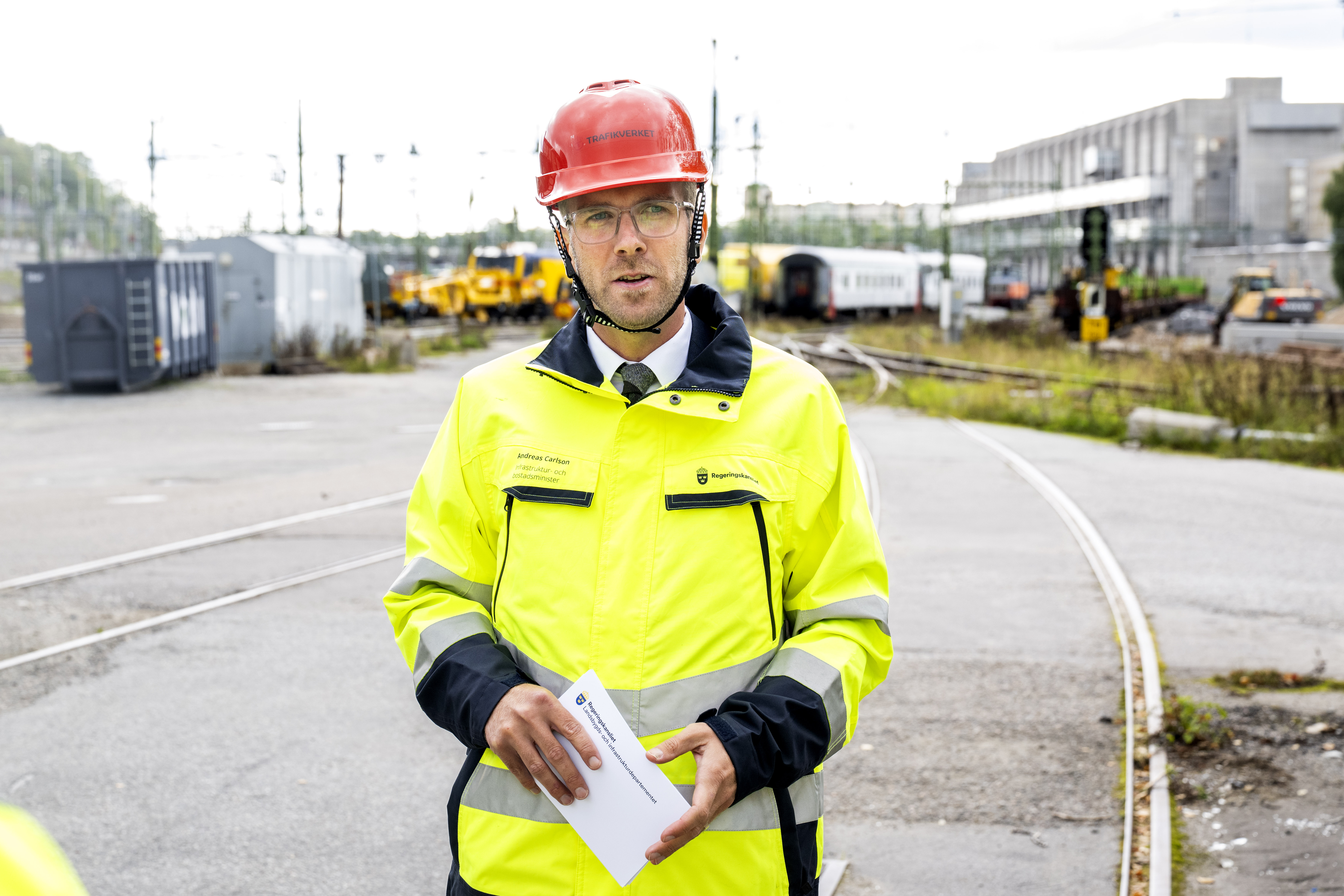 En person med röd skyddshjälm och gul varseljacka står vid järnvägsspår och håller papper på en industriplats.