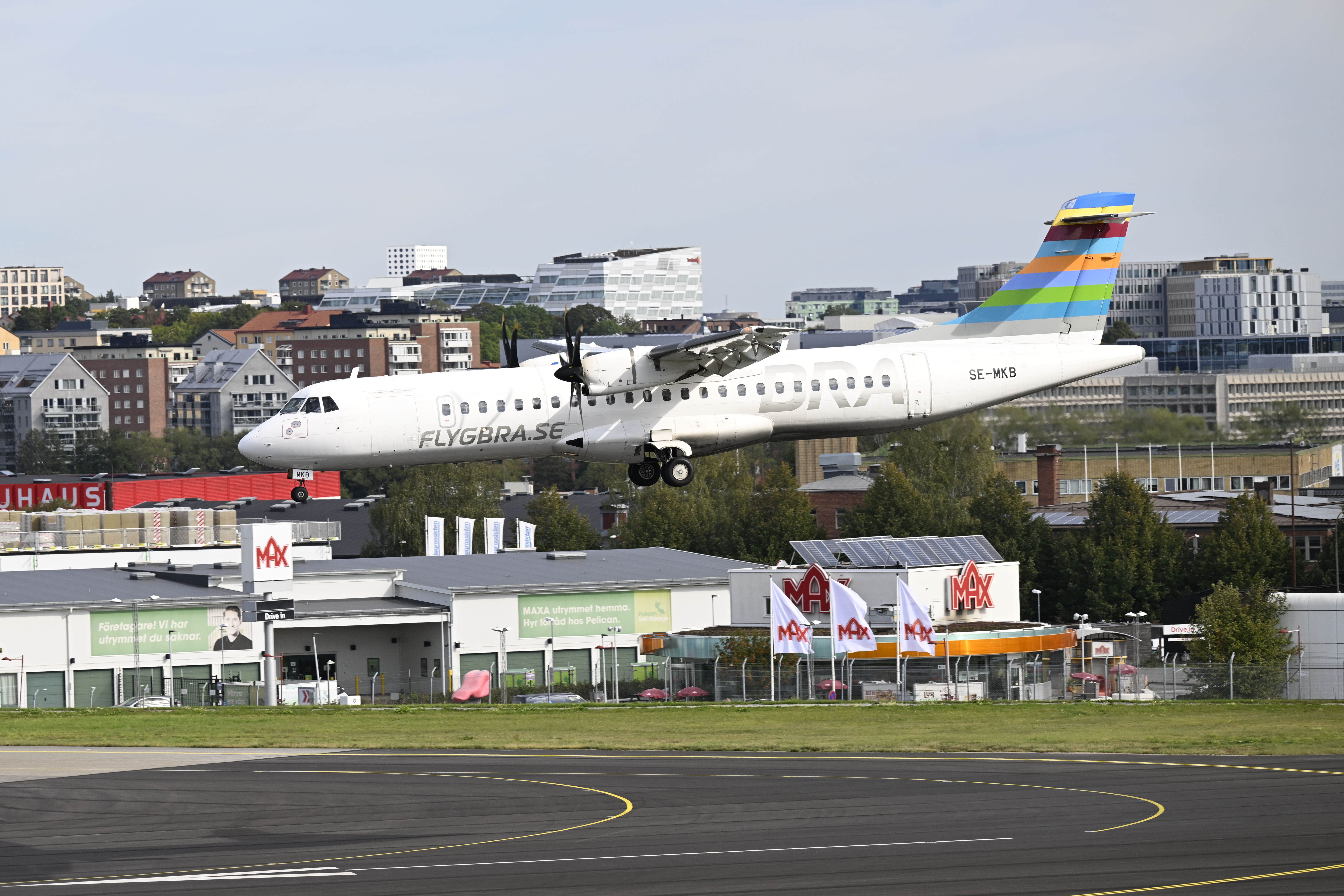 Ett Flygbra.se turbopropflygplan med färgad stjärt landar på en stadsflygplats, med byggnader och butiker i bakgrunden.